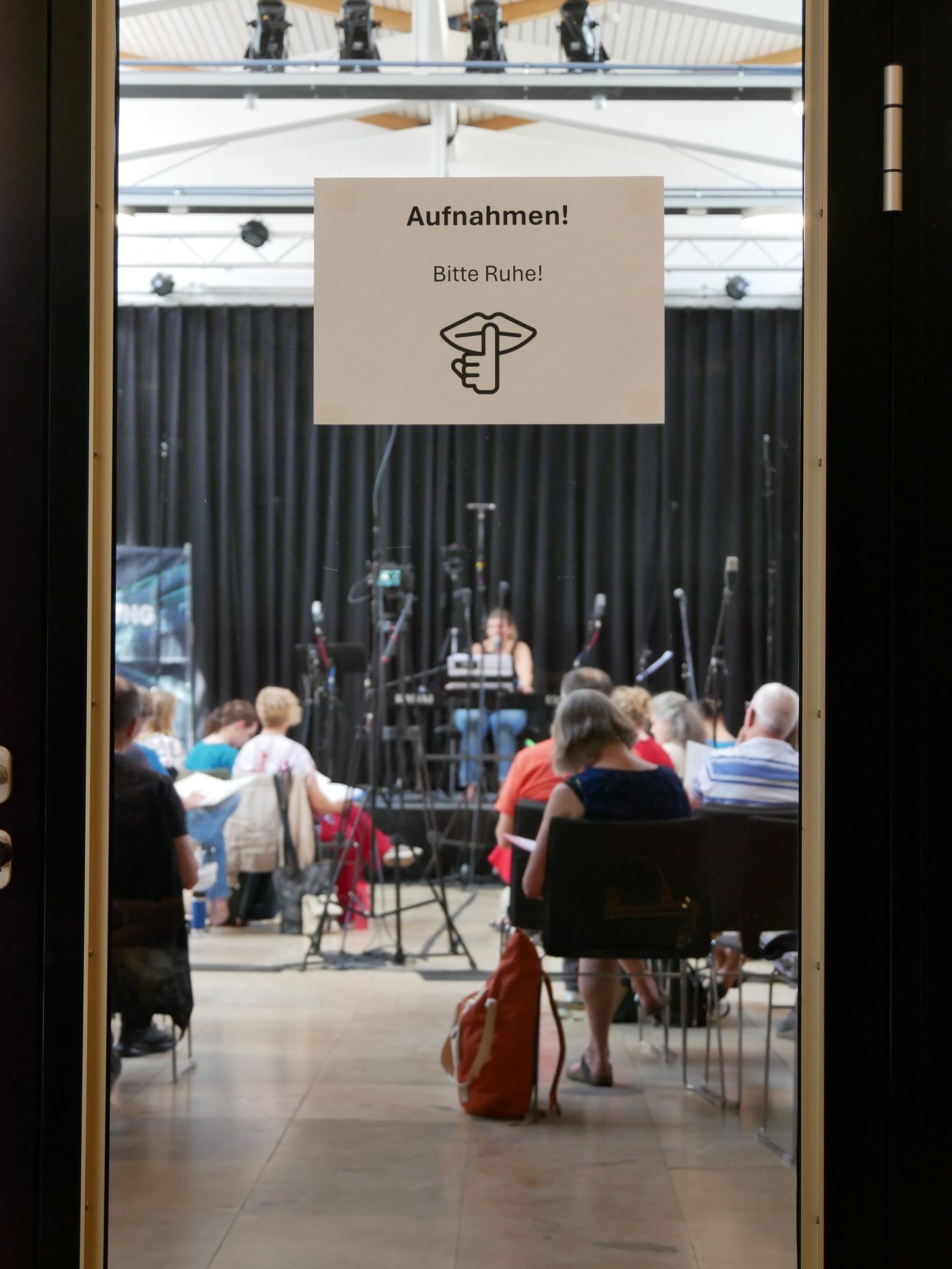 A "Recordings! Please be quiet!" sign on a glass door, showing people and recording equipment inside.