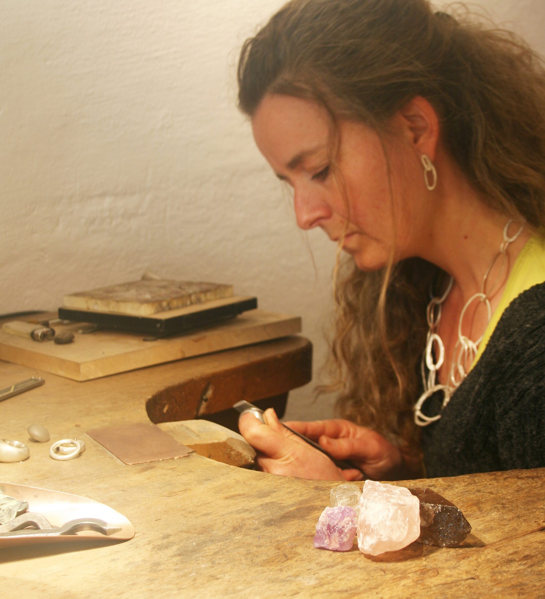 Woman jeweler working at a bench with tools and raw gemstones.