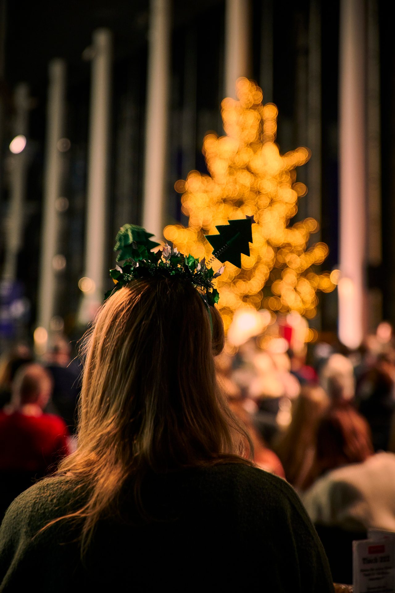 Person with Christmas tree headband facing blurred glowing Christmas tree lights.