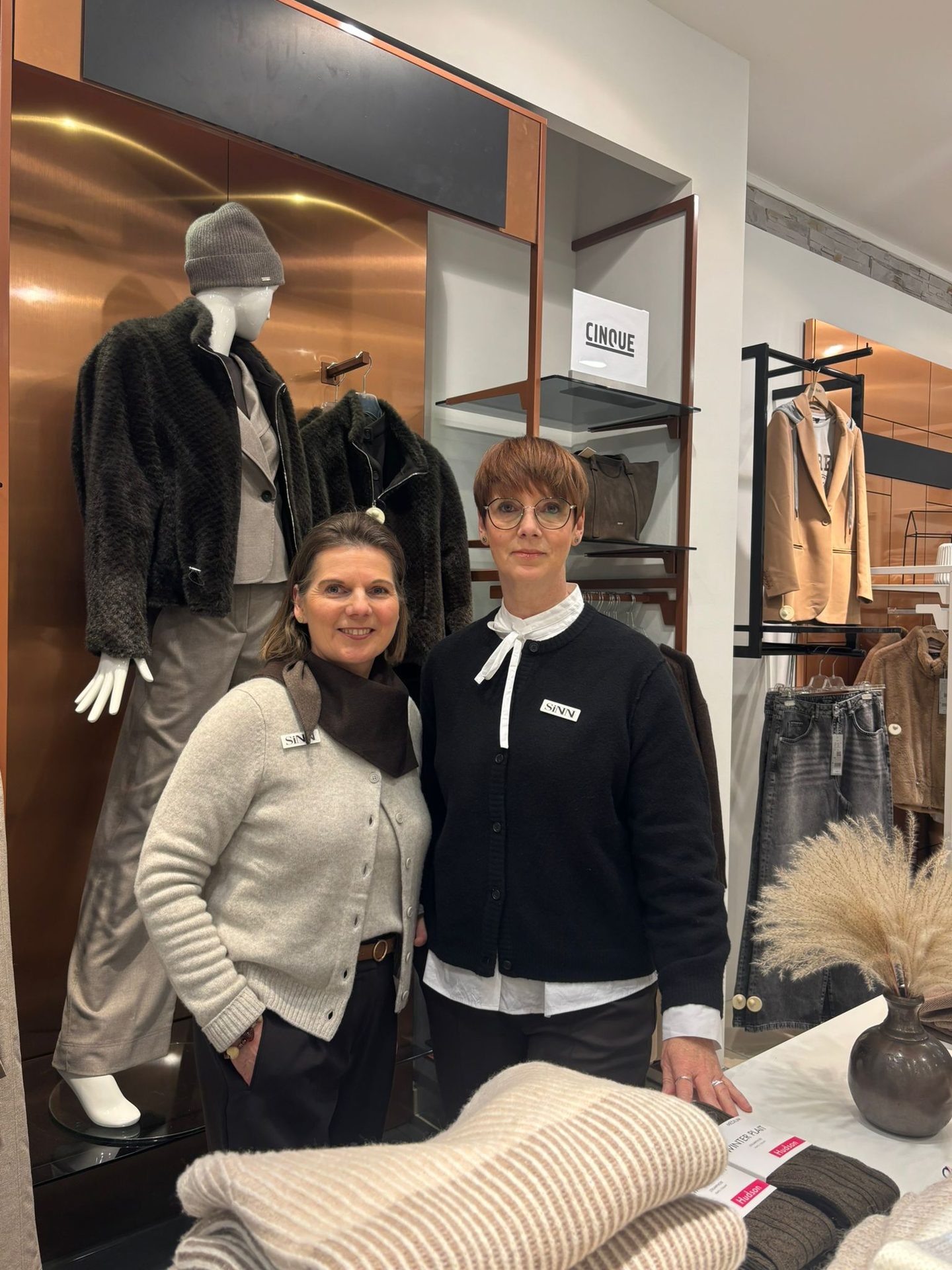 Two women stand in a clothing store with mannequins and clothing displays in the background.
