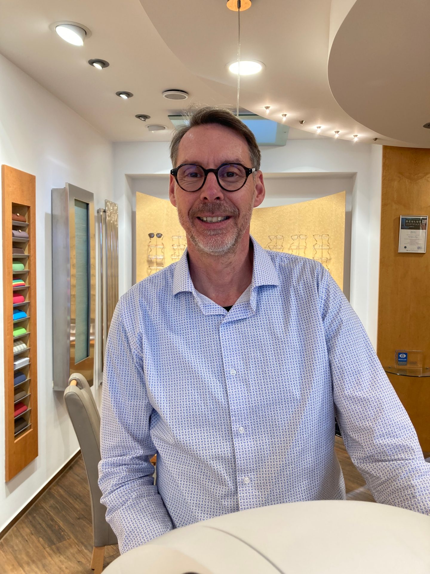 Smiling man with glasses and patterned shirt in an optician's shop.