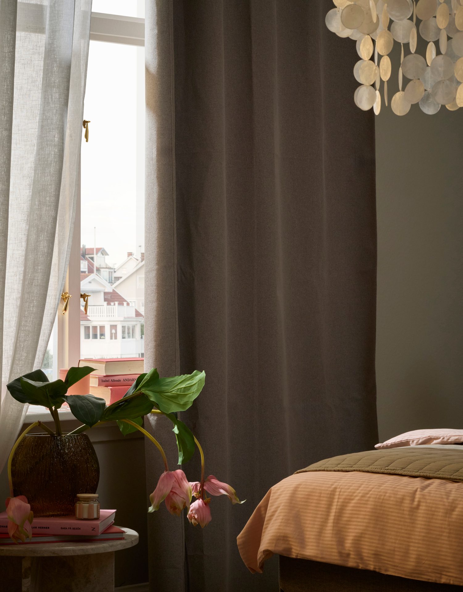 Cozy bedroom with light and dark curtains, a potted plant with pink flowers, books, and a bed.