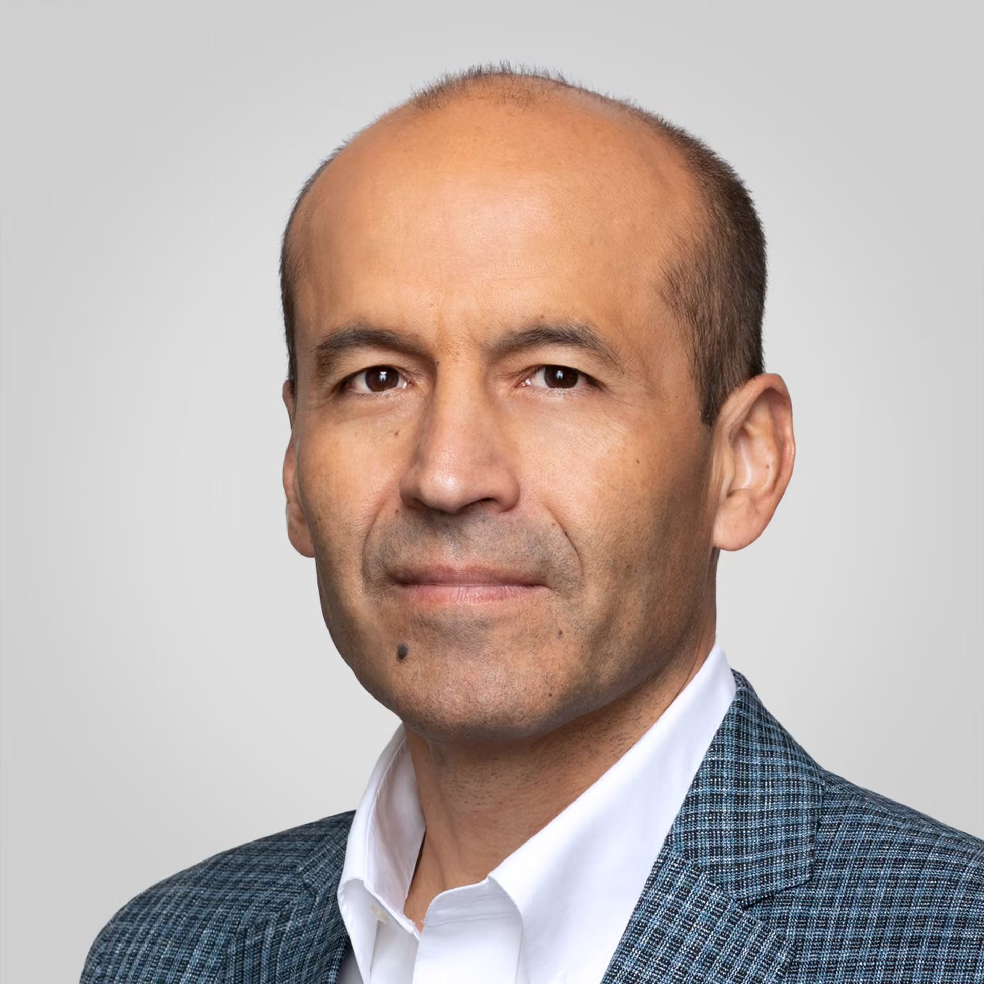 Man with dark eyes, receding hairline, wearing white shirt and blue plaid blazer.