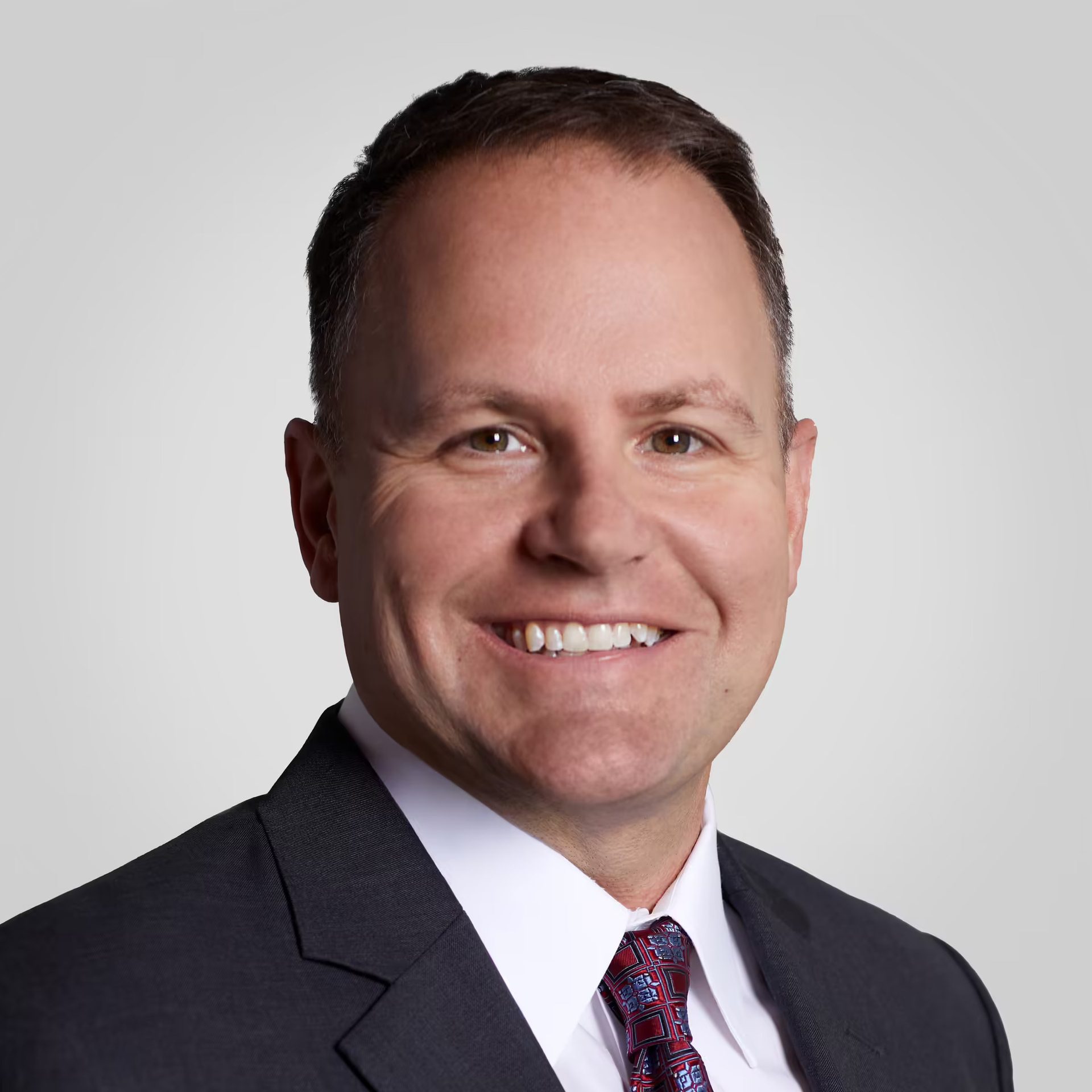 Professional headshot of a smiling man in a dark suit and red patterned tie.