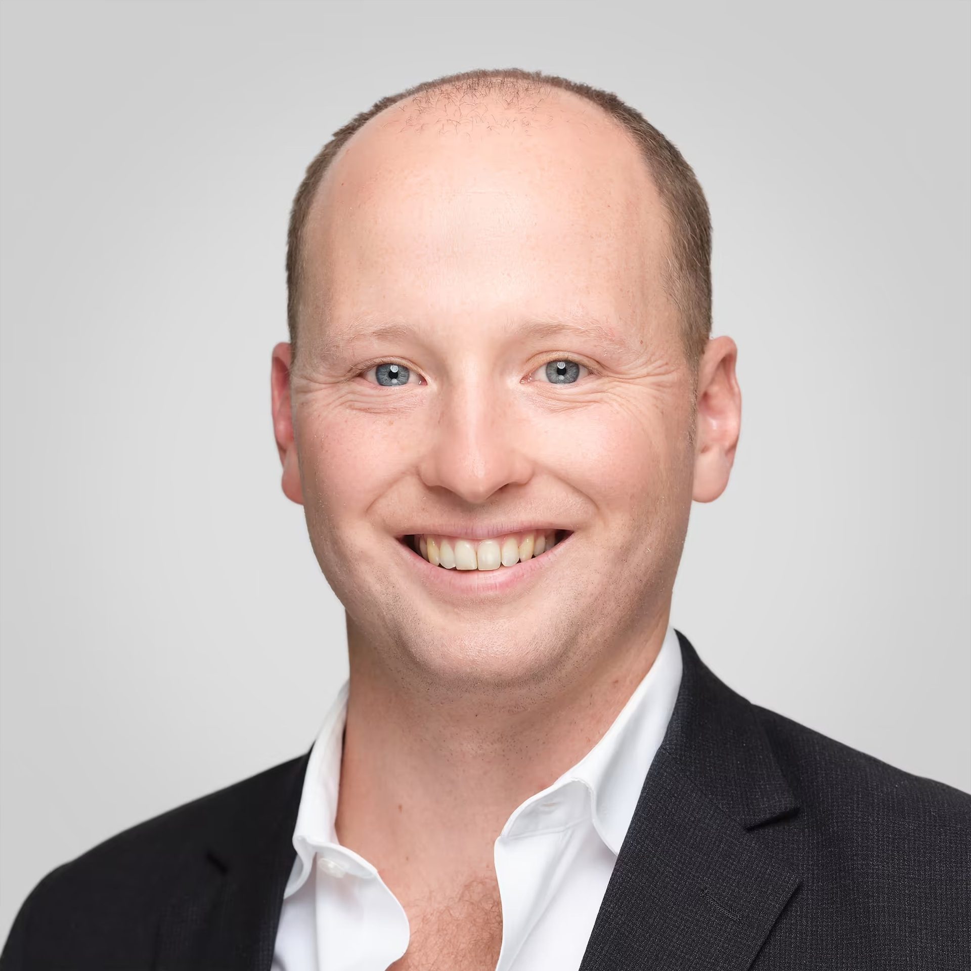 Headshot of a smiling man with blue eyes, wearing a black suit and white shirt.