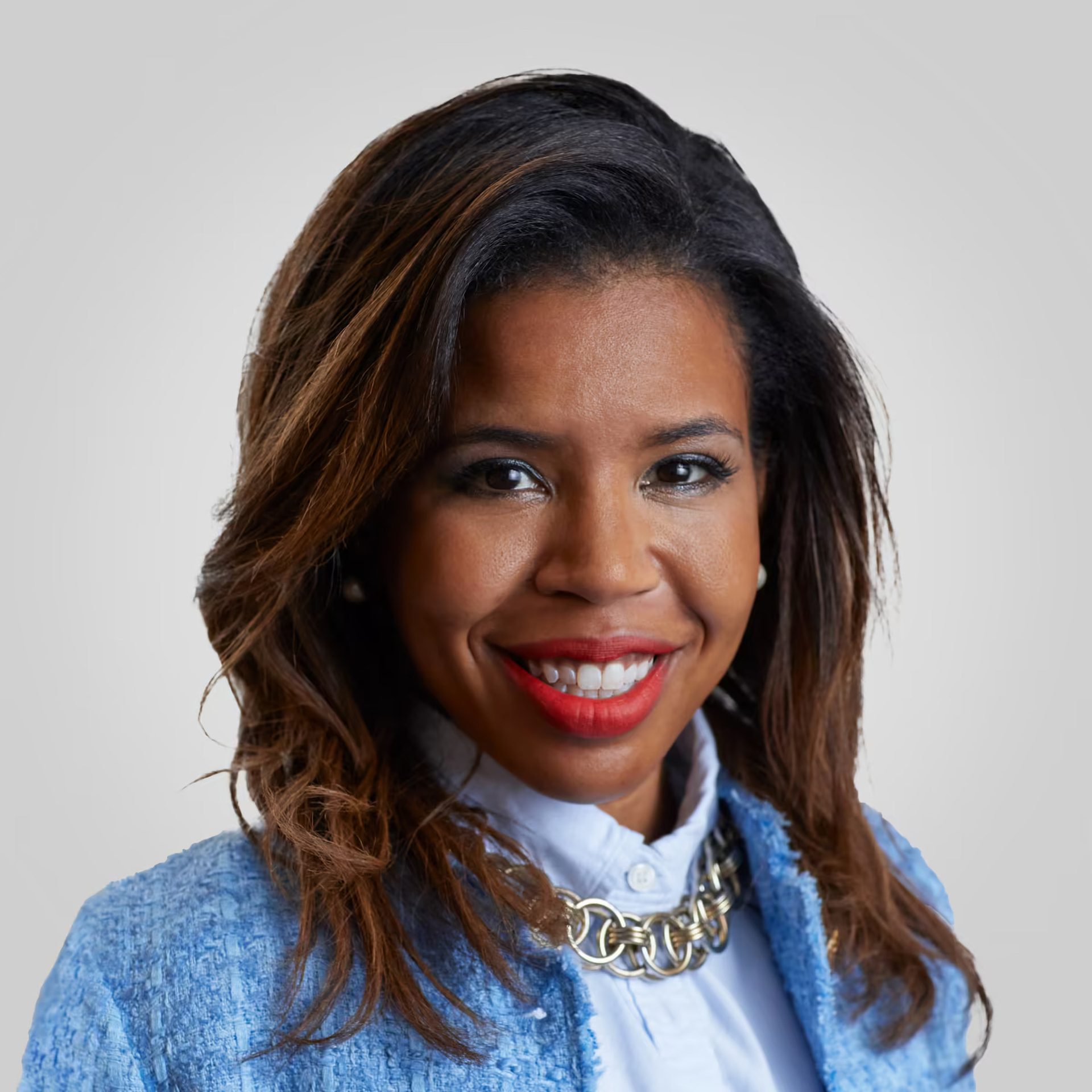 Headshot of a smiling woman with dark hair, red lipstick, light blue jacket, and a chain necklace.