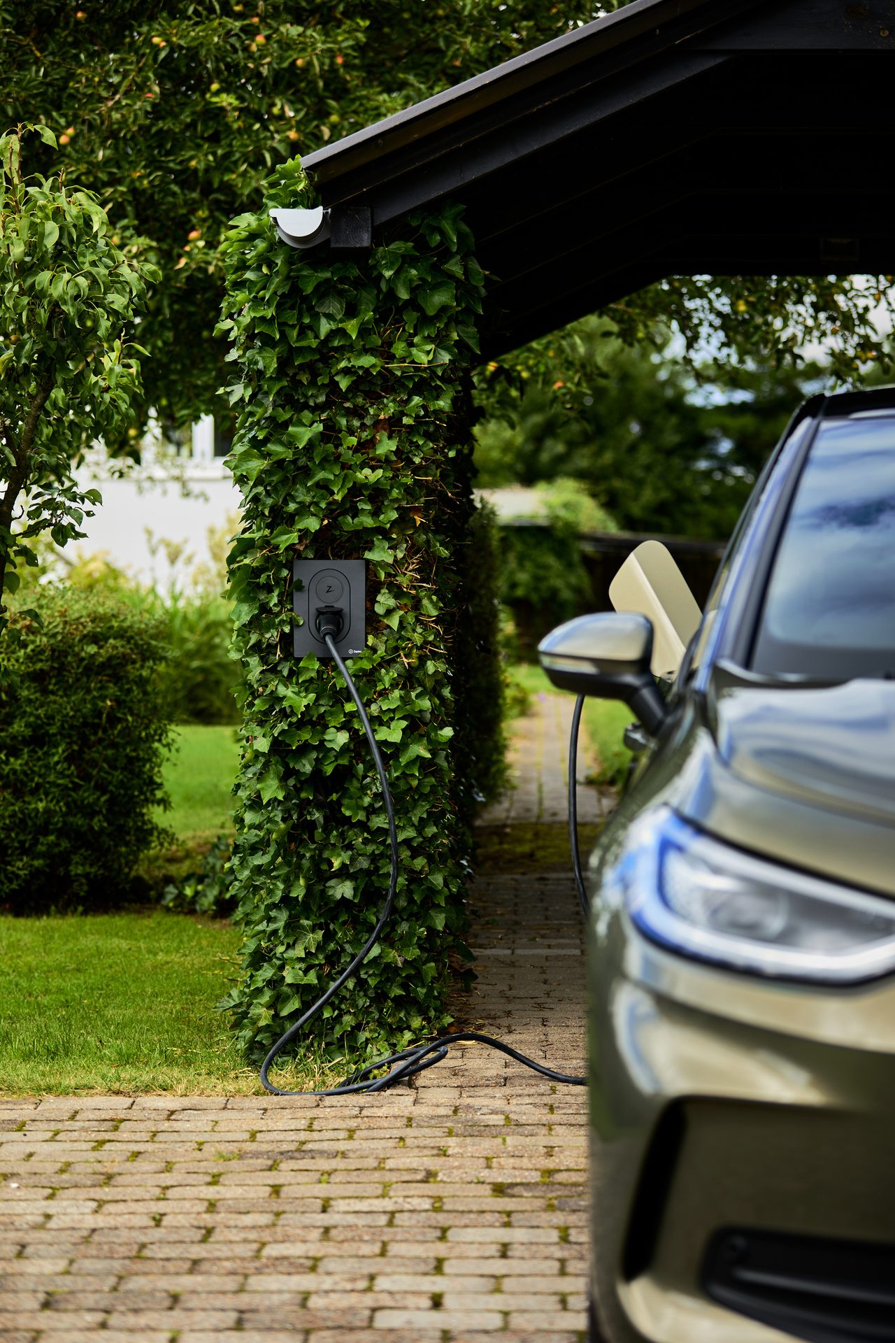 Electric car charging from a wall charger on an ivy-covered pillar next to a brick driveway.