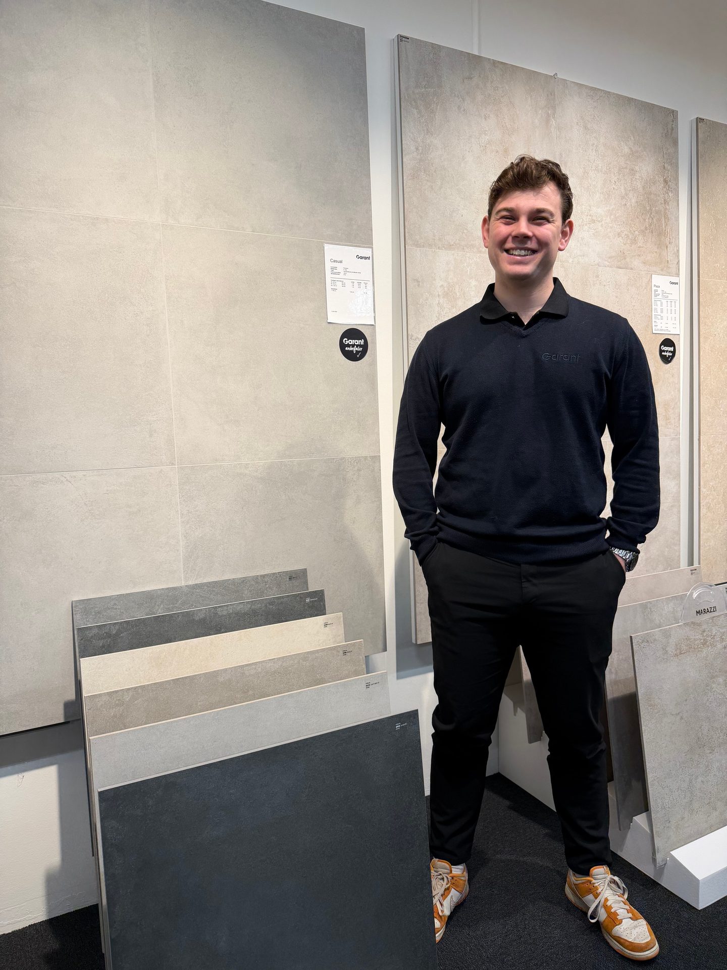 A smiling man in a dark sweater stands amidst various tile displays in a showroom.