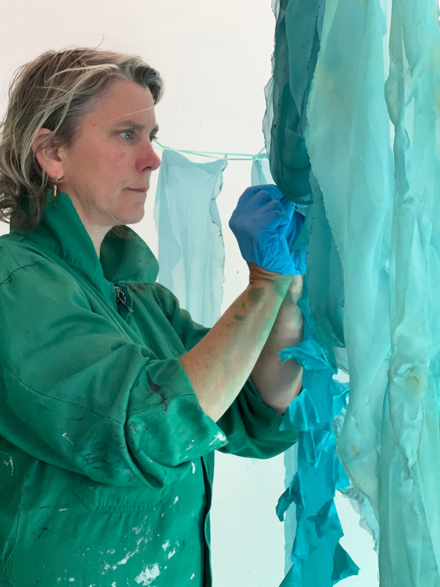 Woman in green overalls and blue gloves manipulates light blue fabric, likely dyeing or crafting.