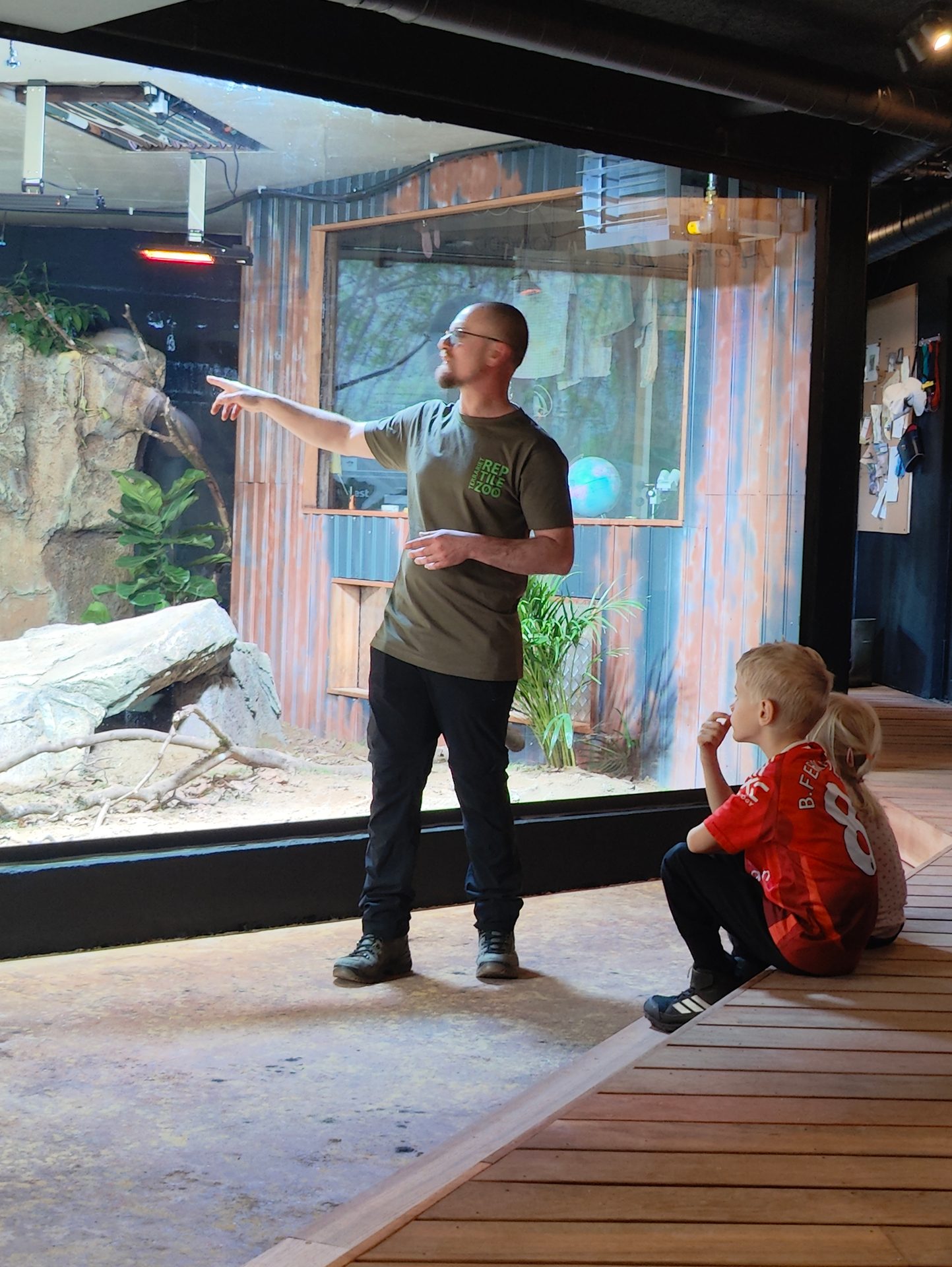 A man in a green t-shirt points towards an enclosure at a reptile zoo, as two children watch intently.