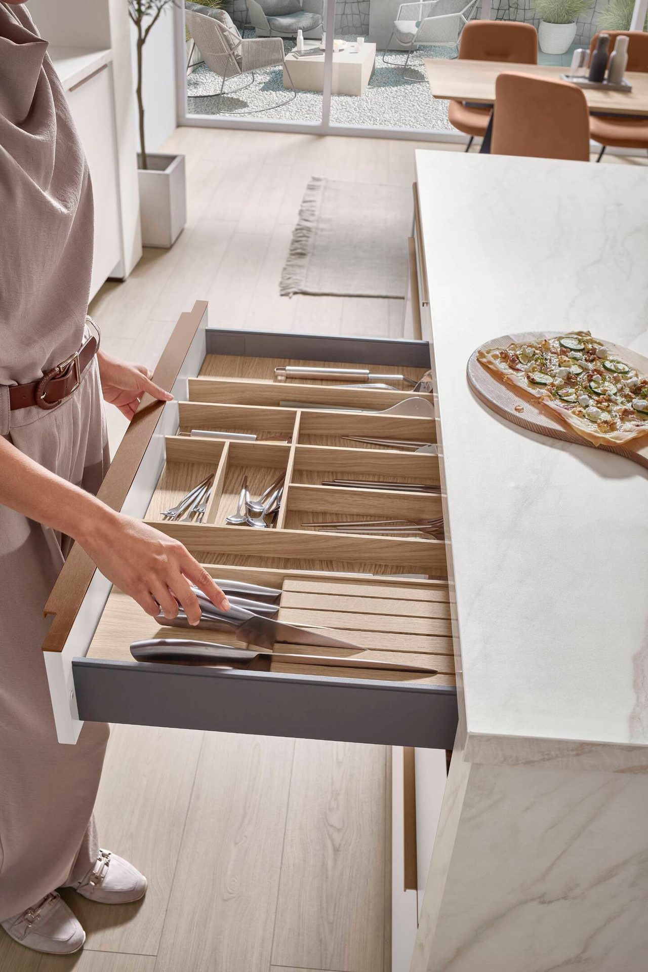Woman opening a kitchen drawer with organized cutlery, revealing a modern kitchen and outdoor patio.