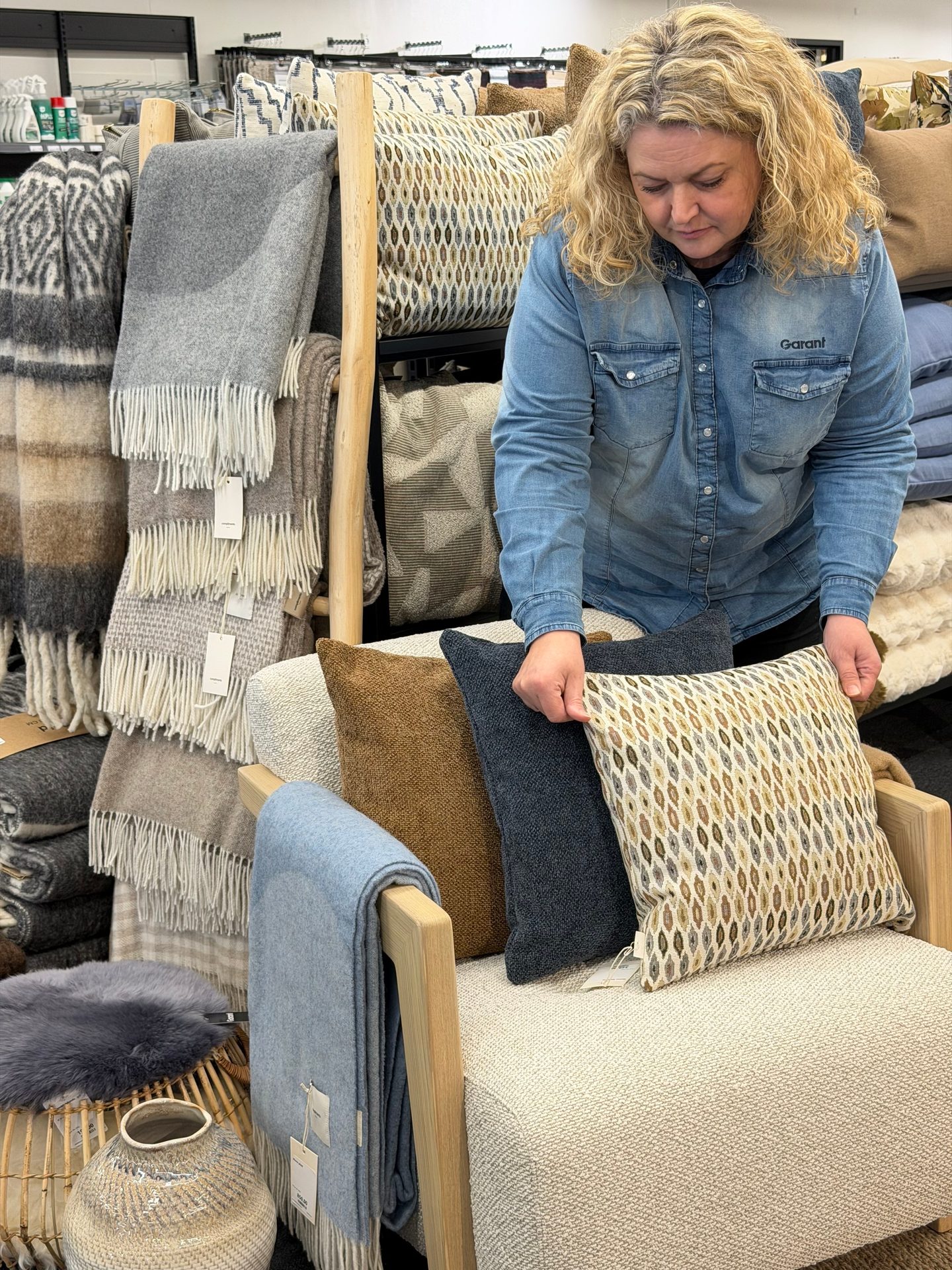 A woman in a denim shirt arranges decorative pillows on a chair in a home decor store, surrounded by blankets.