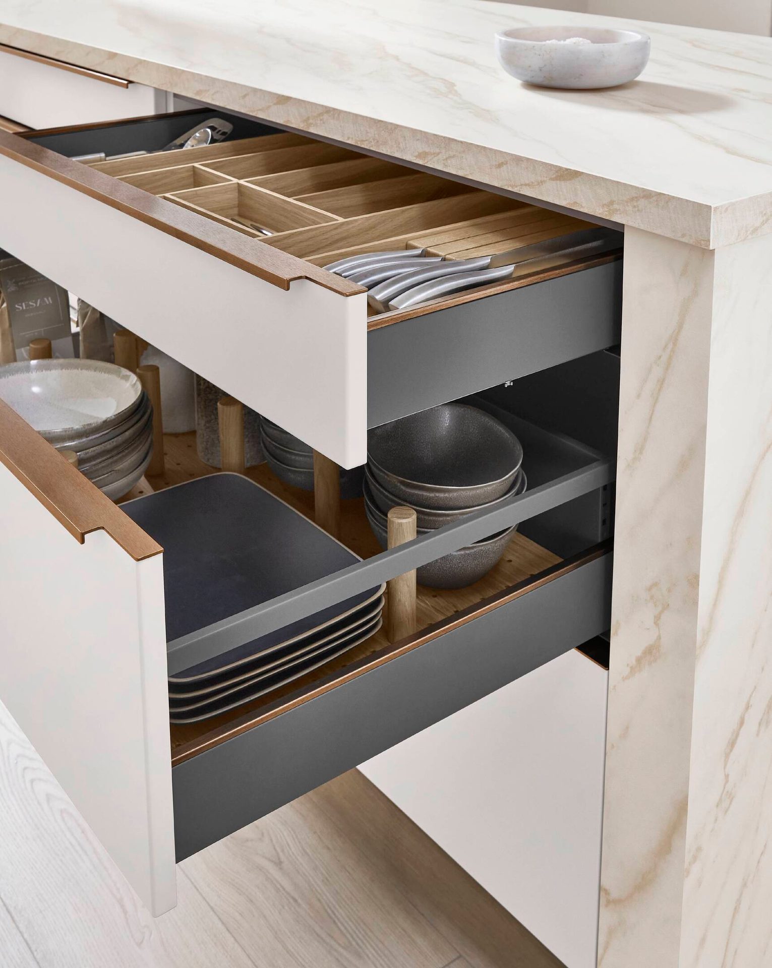 Open kitchen drawers revealing organized cutlery, plates, and bowls, with a marble countertop.