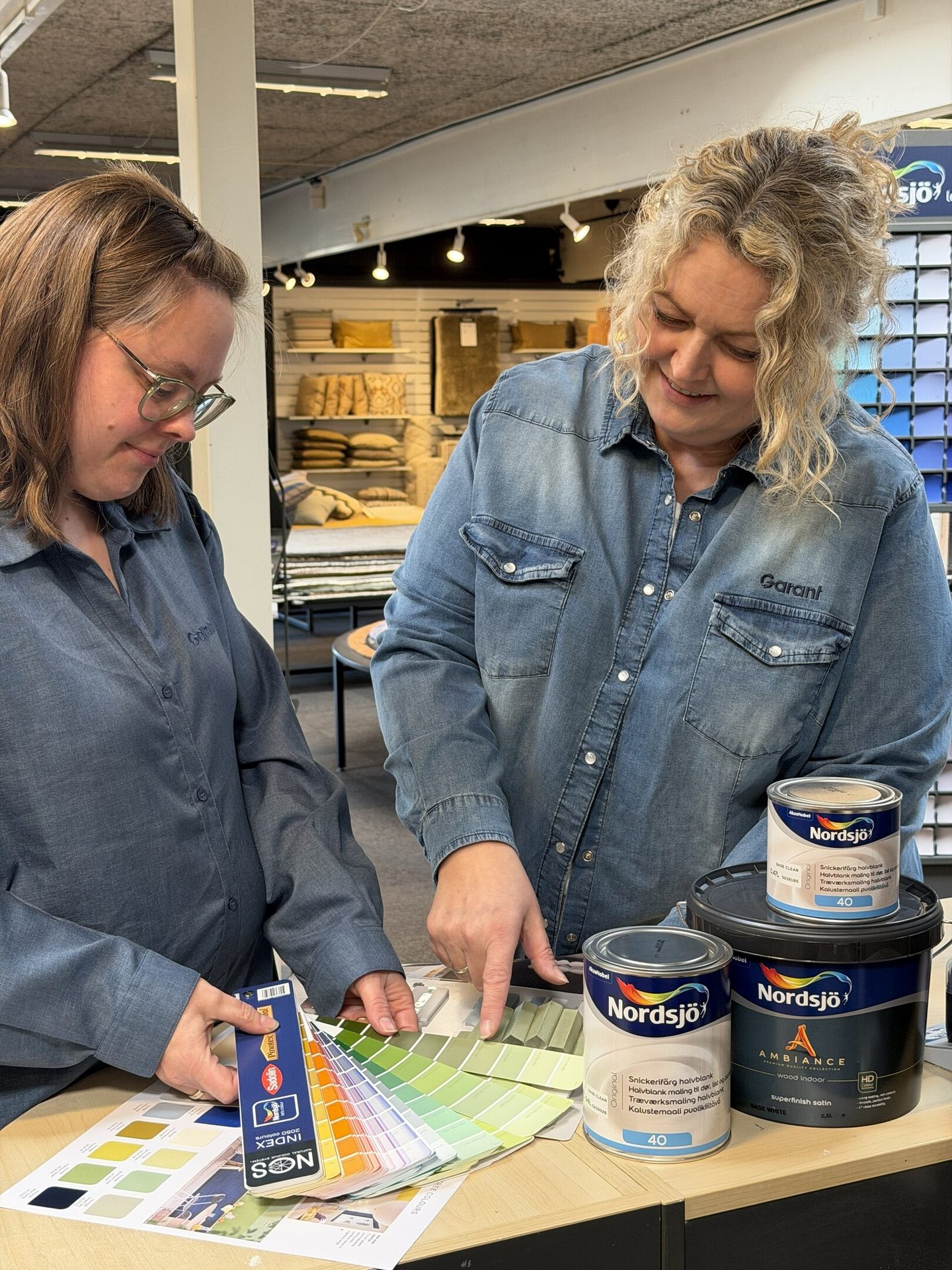 Two women, employees in denim shirts, discuss color choices using swatches and paint cans in a store.