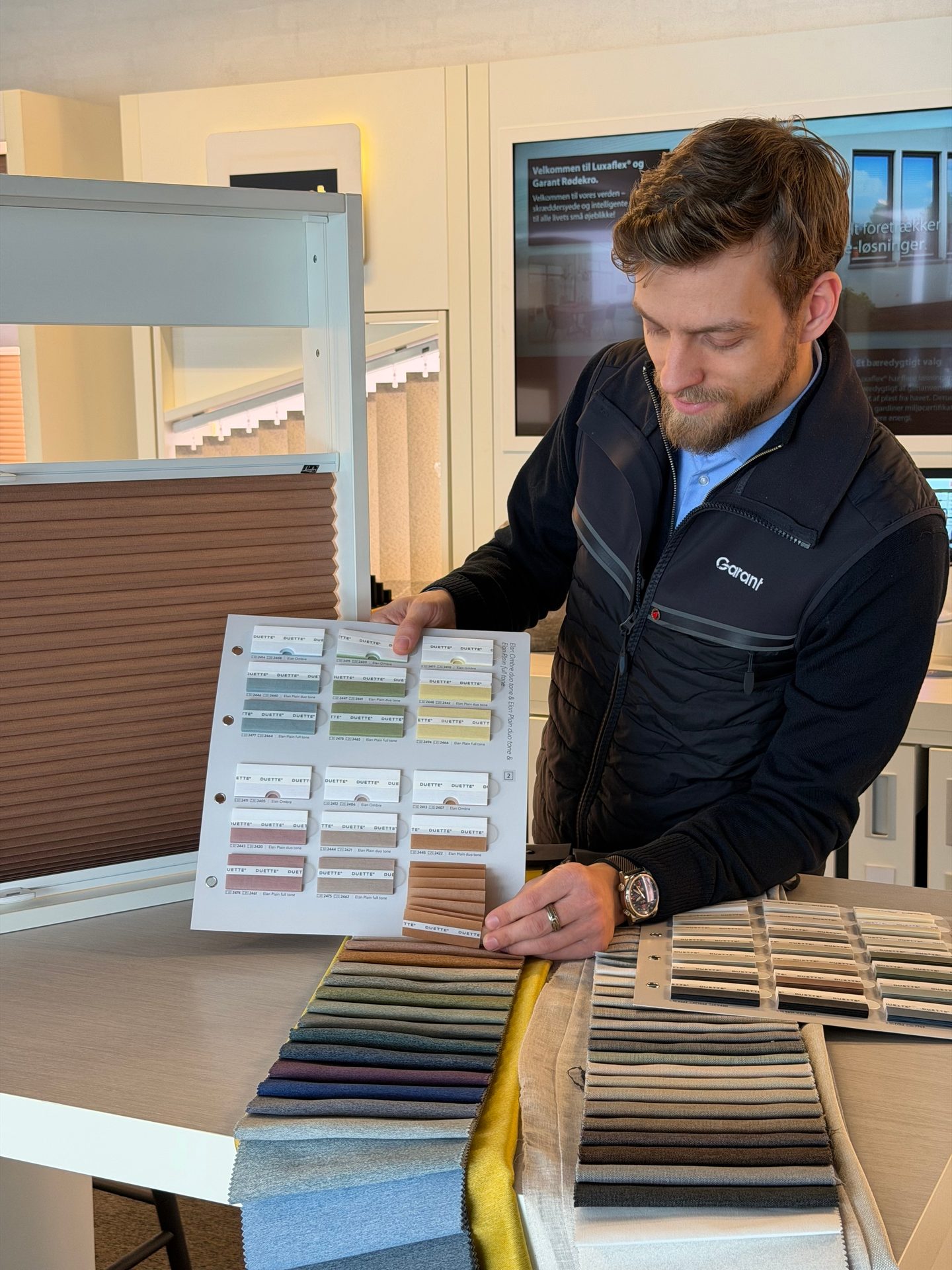 Man in a Garant vest reviews fabric and blind samples on a display card and table for interior design.