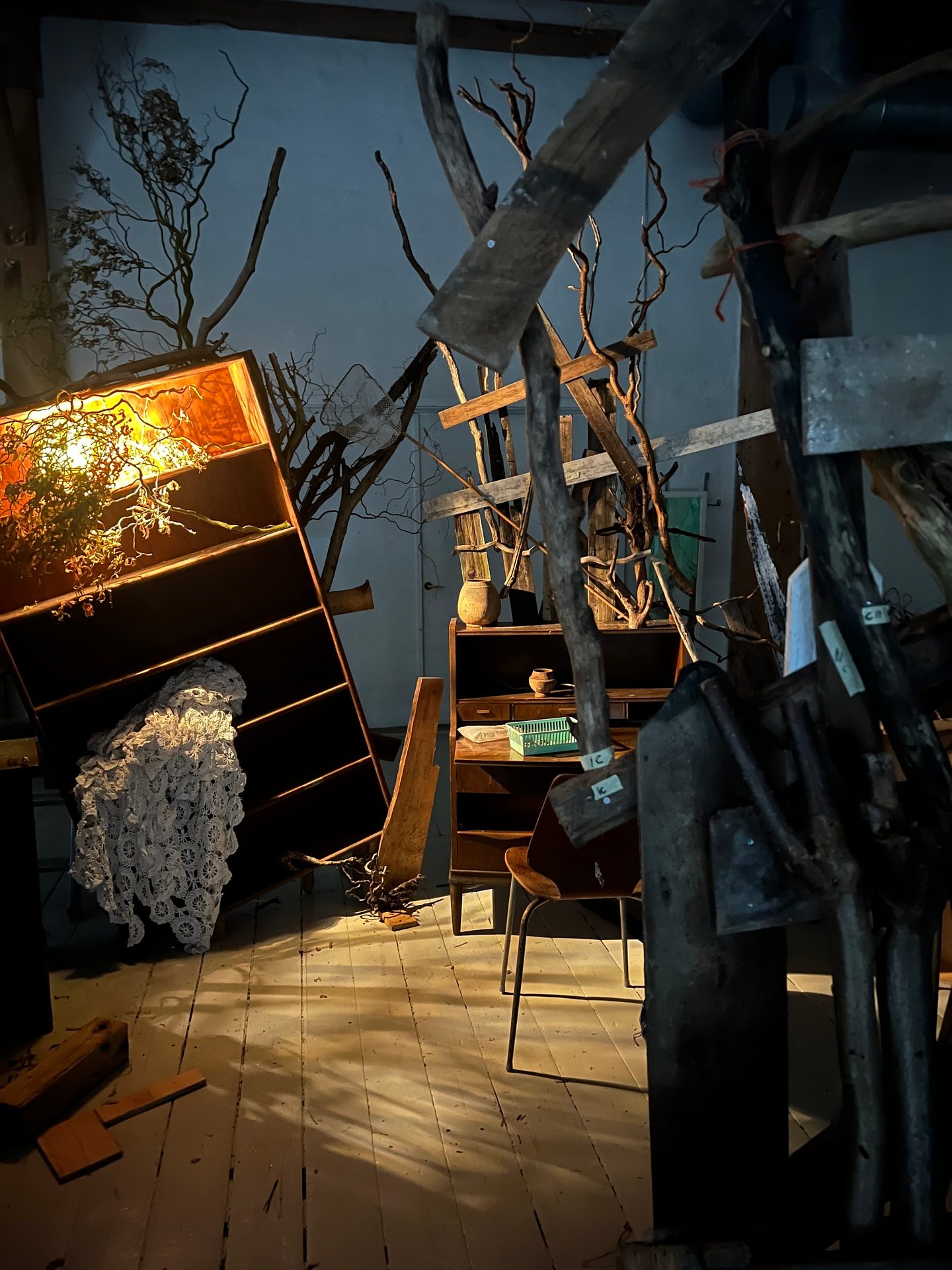 A dimly lit, cluttered room with a chaotic art installation of wood, branches, furniture, and light.