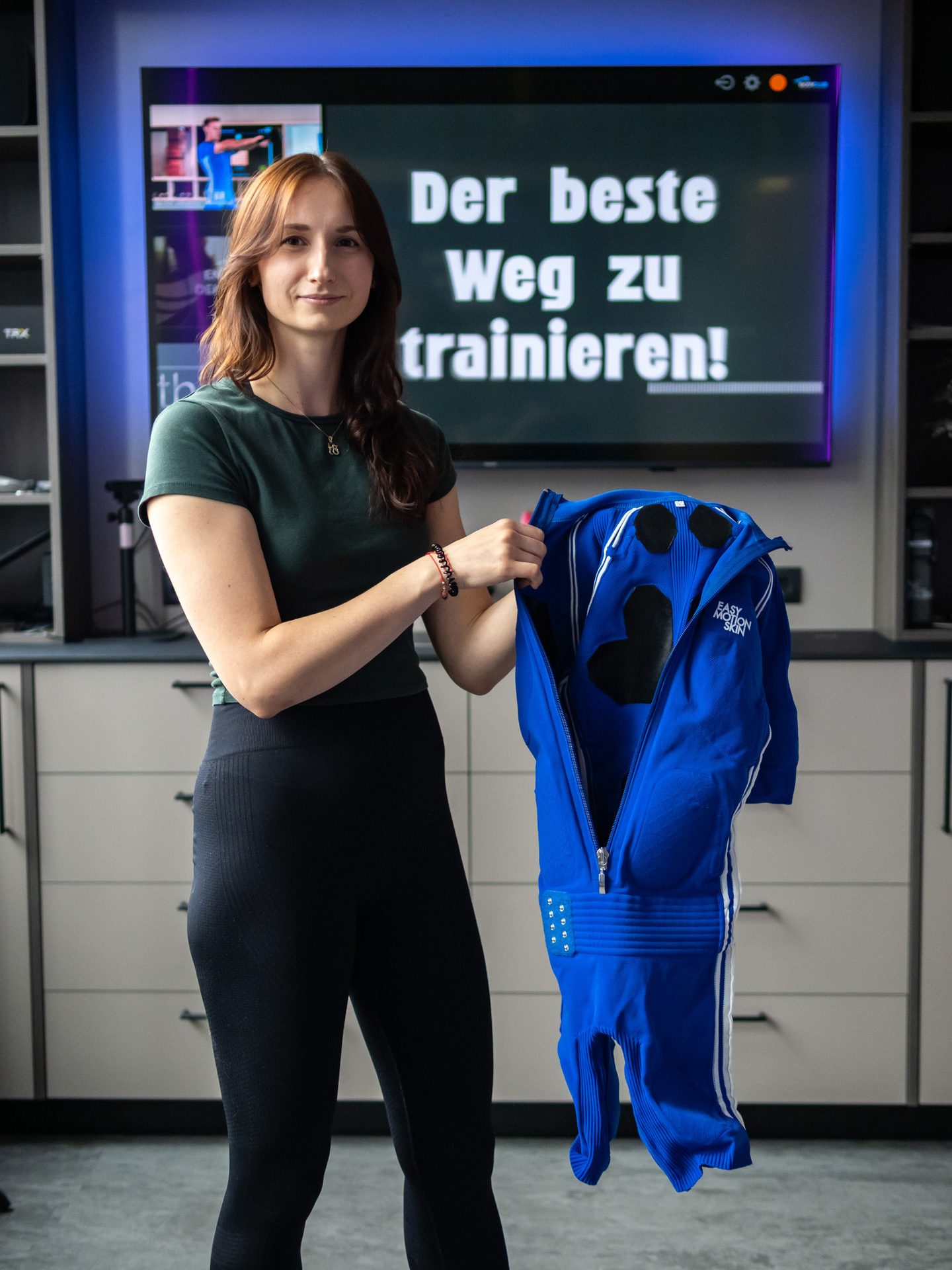 Woman holding a blue EMS training suit in front of a screen displaying German text about training.