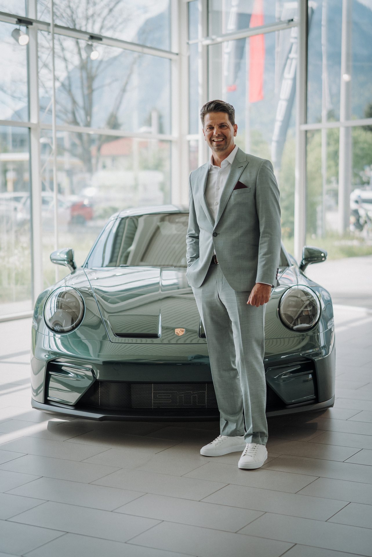 Man in a light gray suit smiling next to a sleek green sports car in a modern showroom.