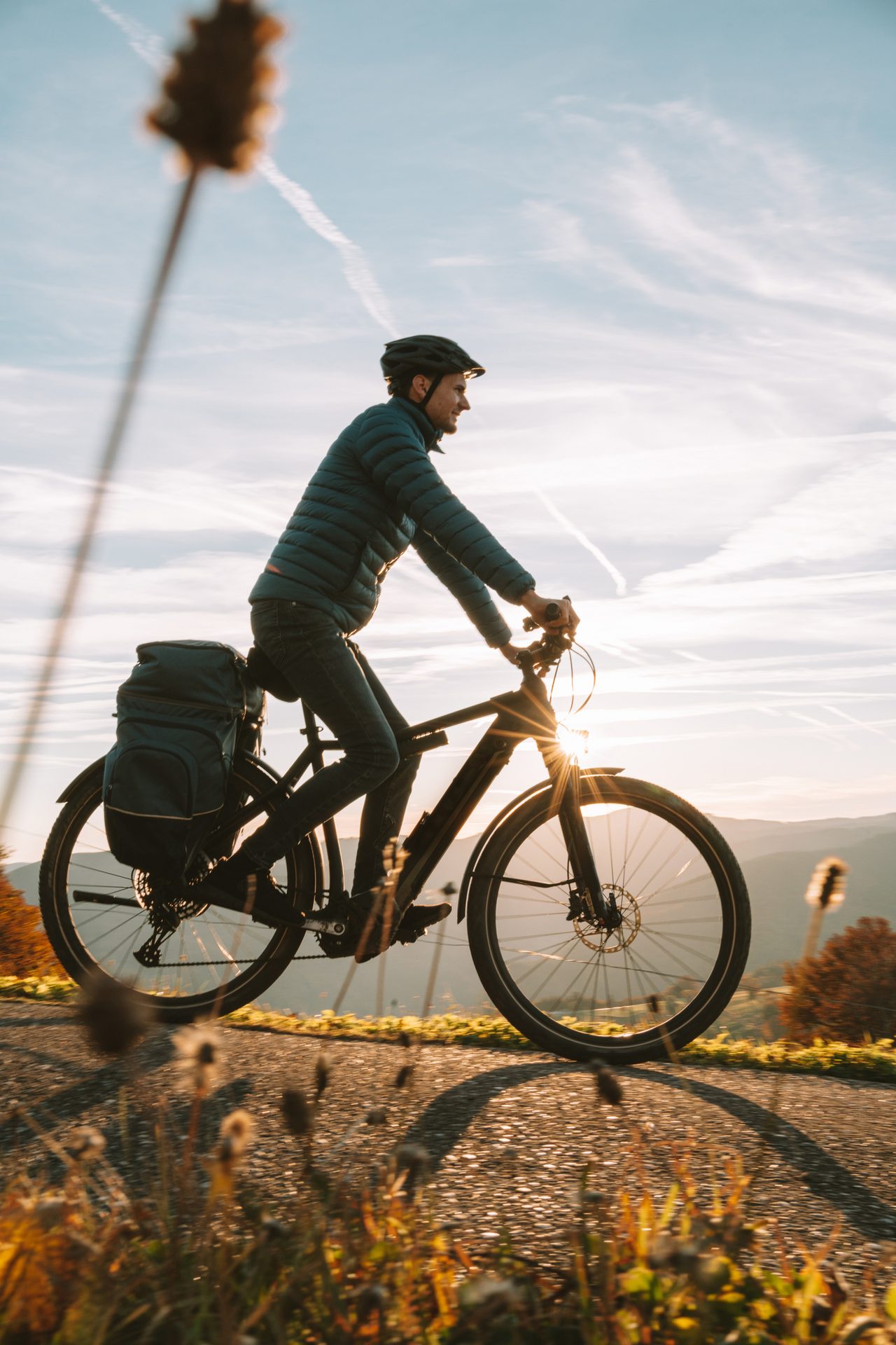 Man in helmet and puffer jacket cycling on a road with panniers, sun flare, and autumn hills at sunset.