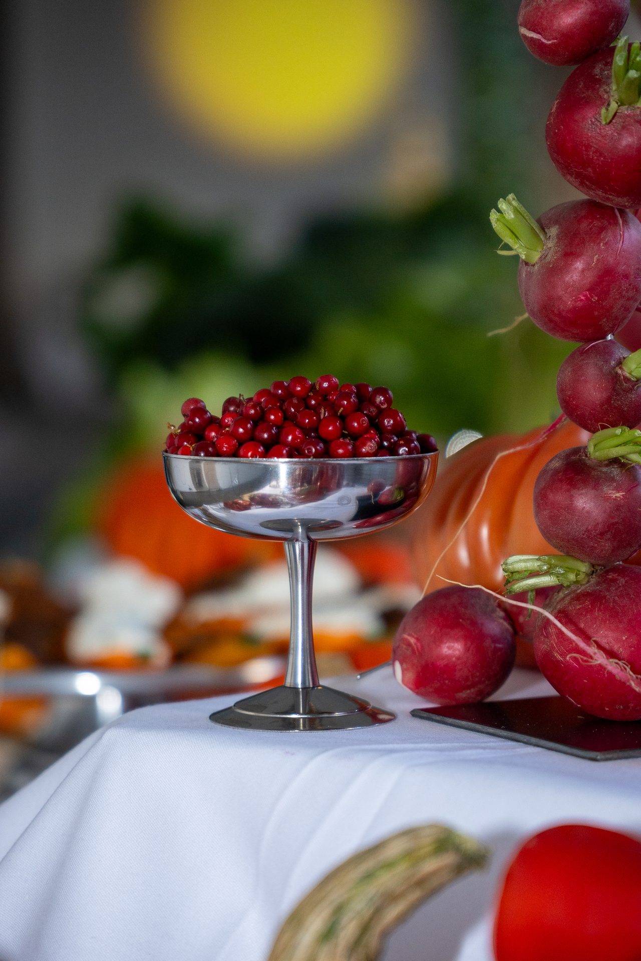 A silver bowl filled with red cranberries next to fresh radishes on a white tablecloth.