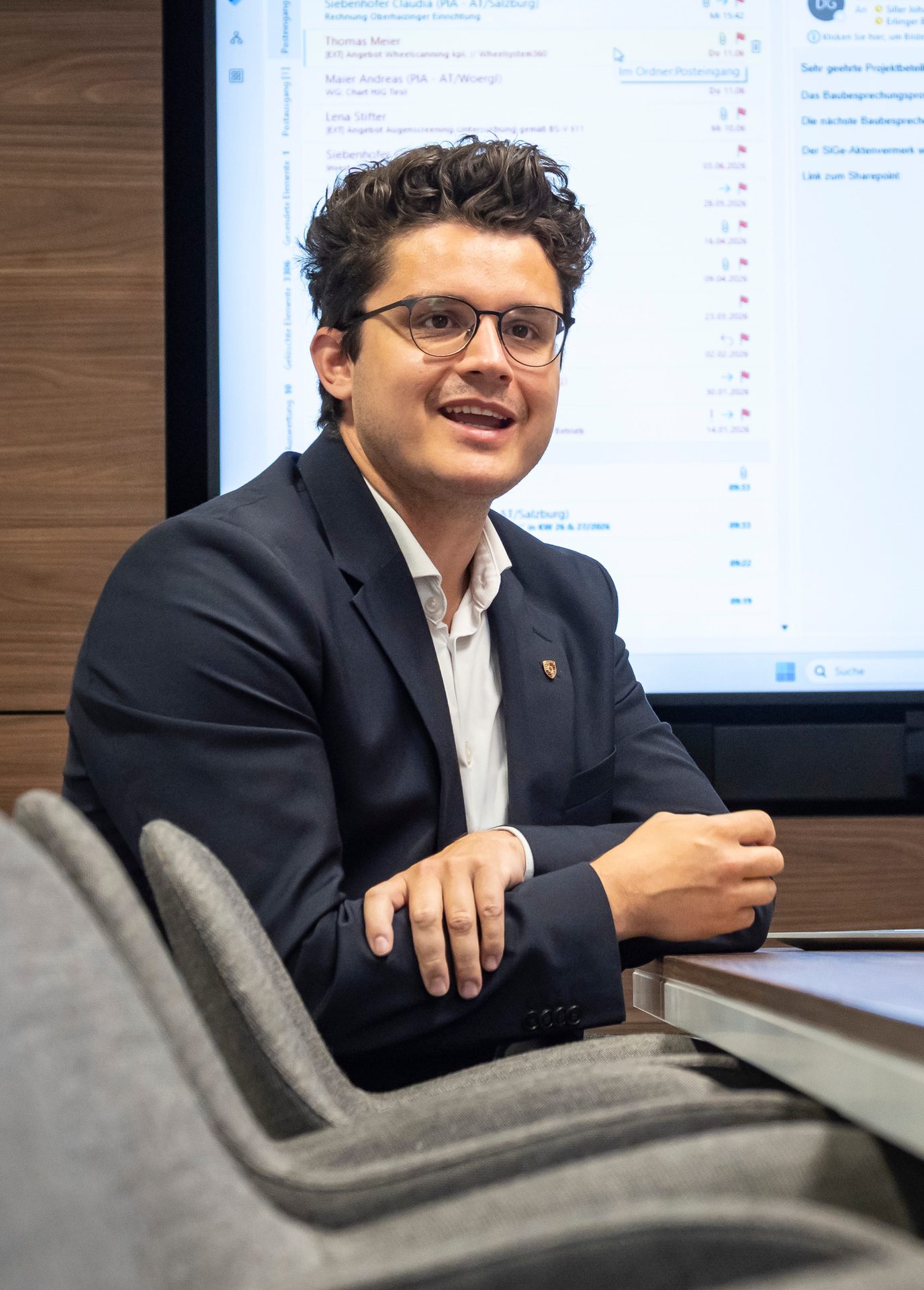 A young man with dark curly hair and glasses, wearing a suit, smiles while seated at a table.