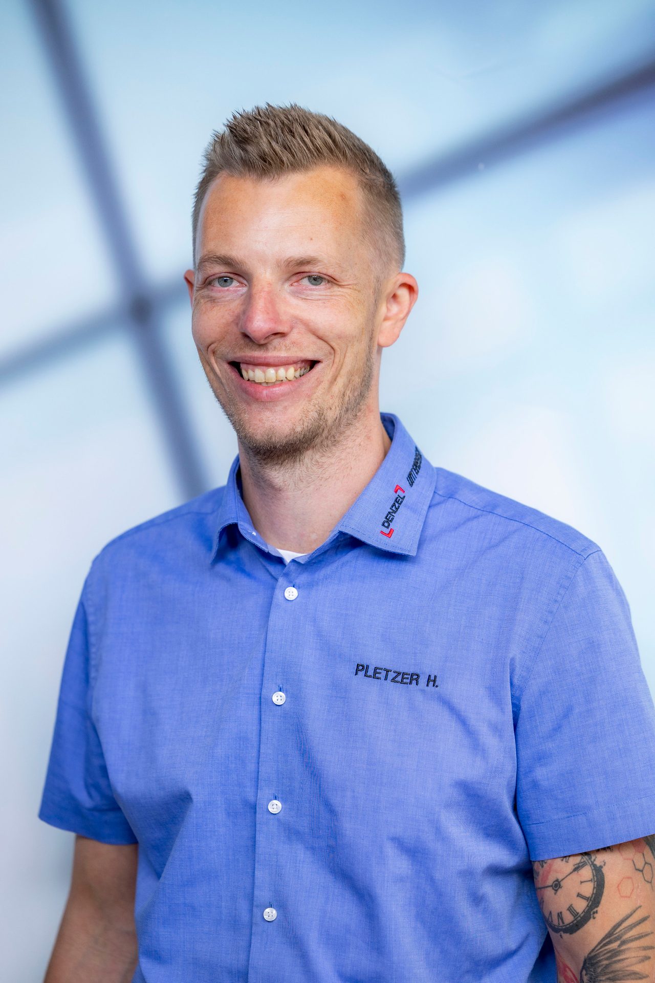Smiling man in a blue shirt with company logos, Pletzer H. and Denzel. He has visible arm tattoos.