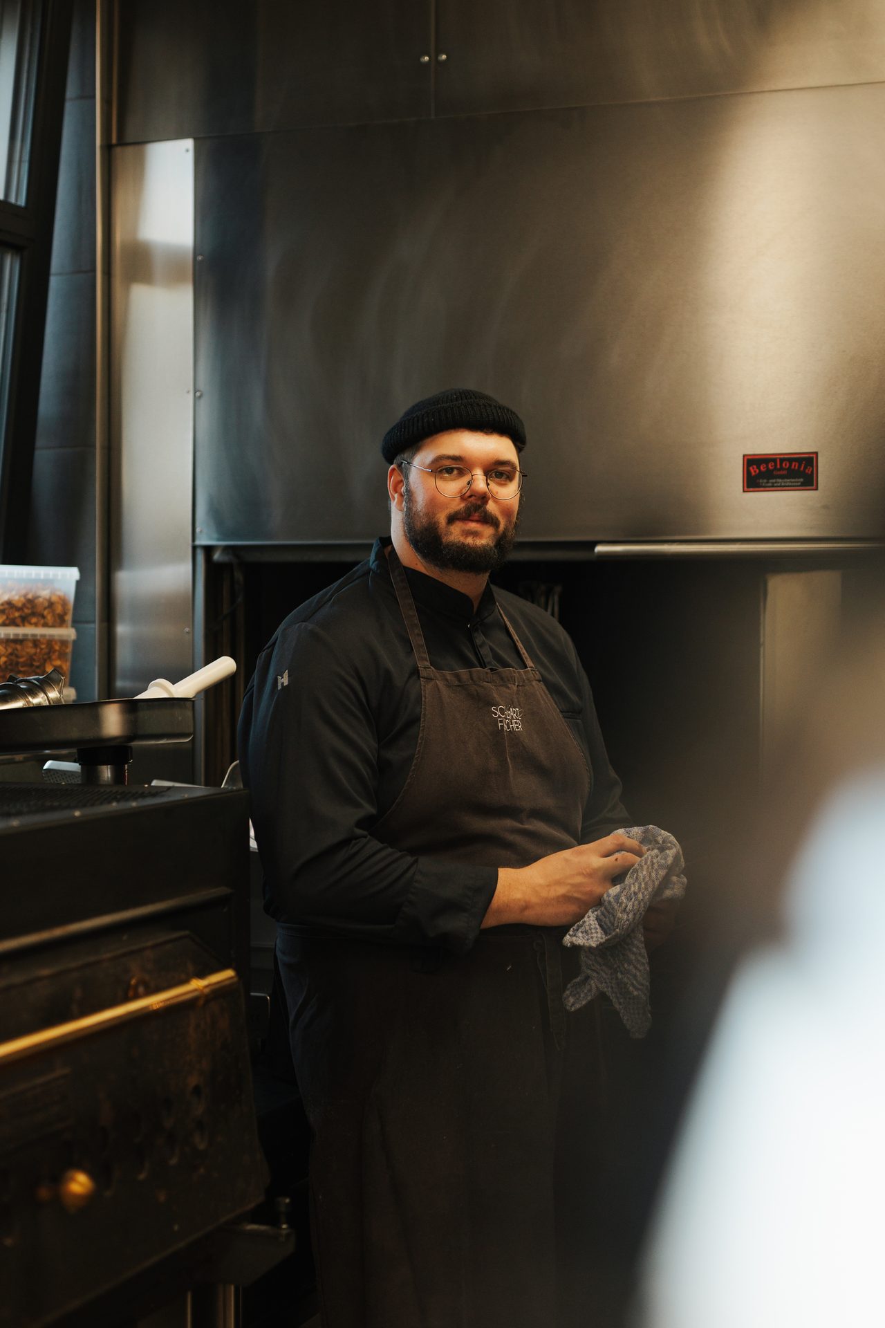 A male chef with a beard and beanie, wearing an apron, stands in a kitchen holding a cloth.