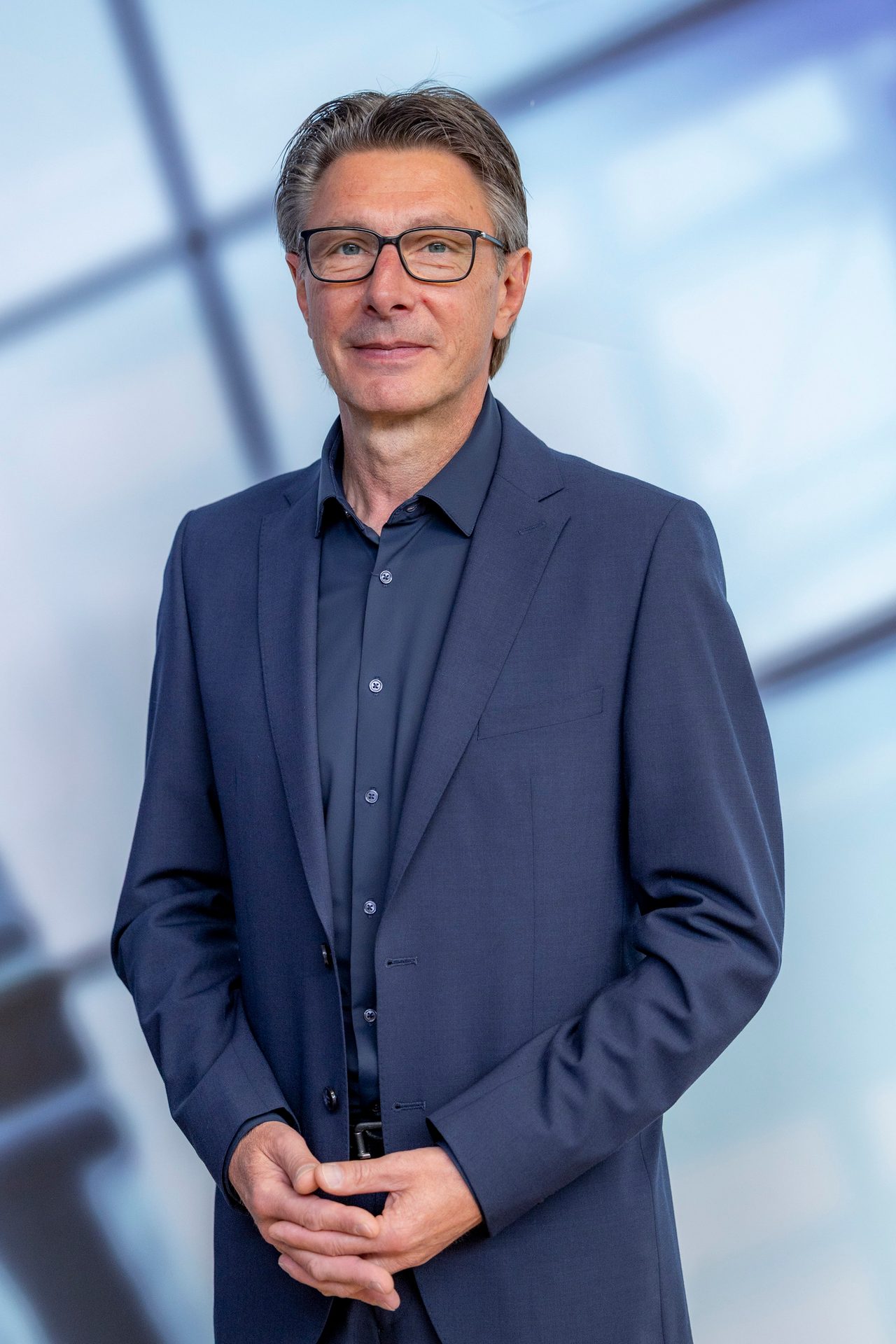 A distinguished man in a dark suit and glasses stands, looking directly at the camera.