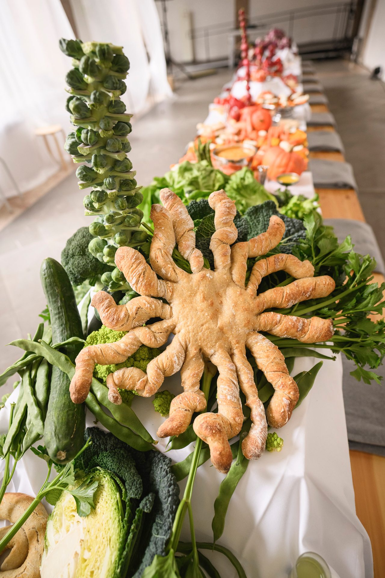 Vibrant buffet with fresh green vegetables, a tall Brussels sprout stalk, and an octopus-shaped bread.