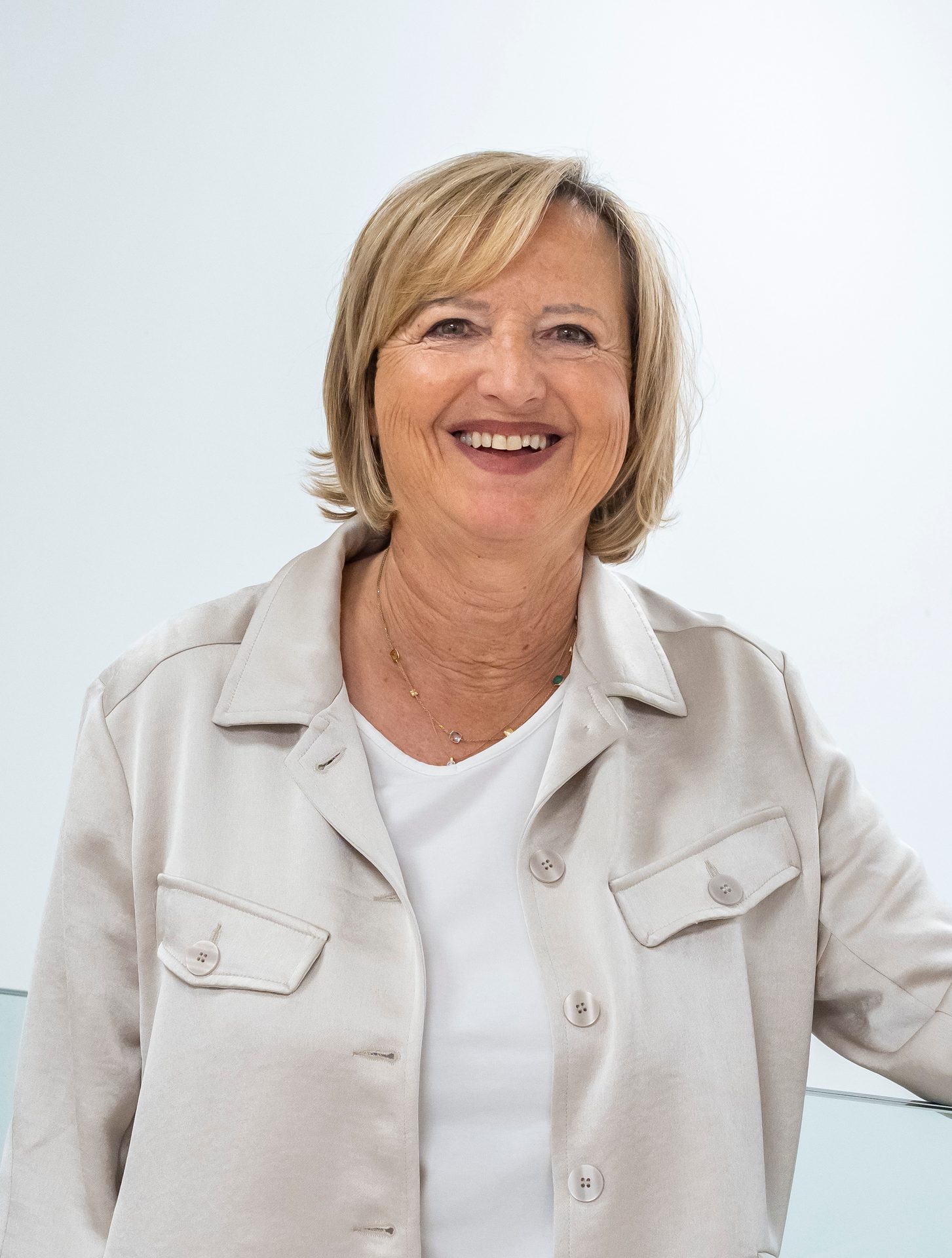 A smiling middle-aged woman with blonde hair, wearing a light jacket and necklace, against a white background.