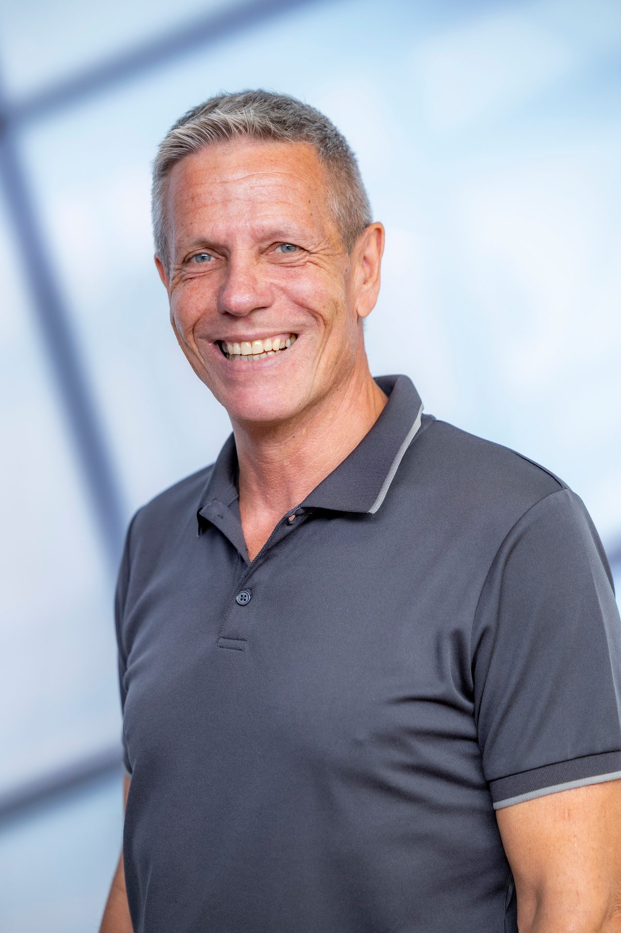 A smiling man with gray hair and a dark gray polo shirt, looking slightly left against a blurred blue background.