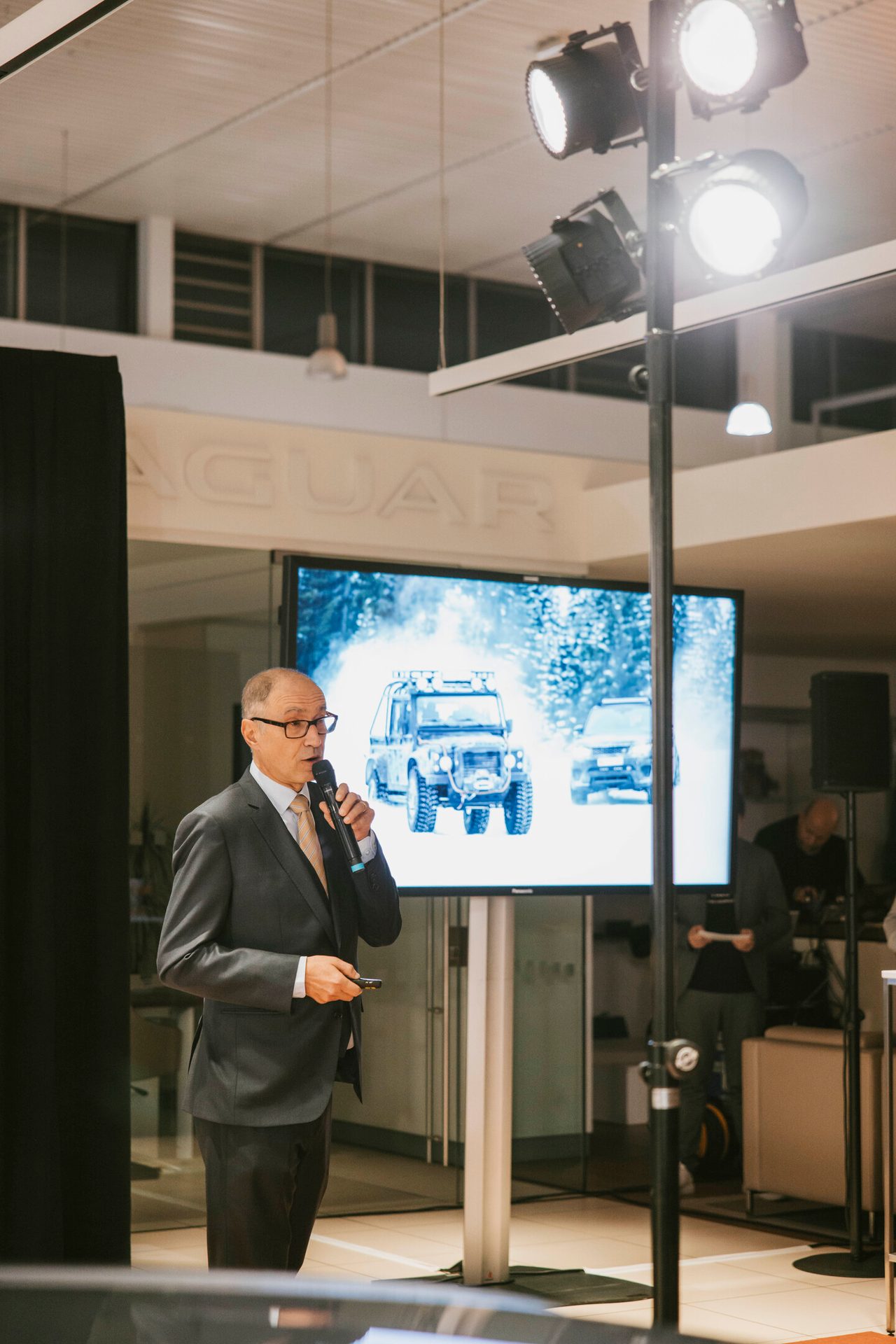 A man in a suit and glasses speaks into a microphone, with SUVs on a screen and a Jaguar logo behind him.