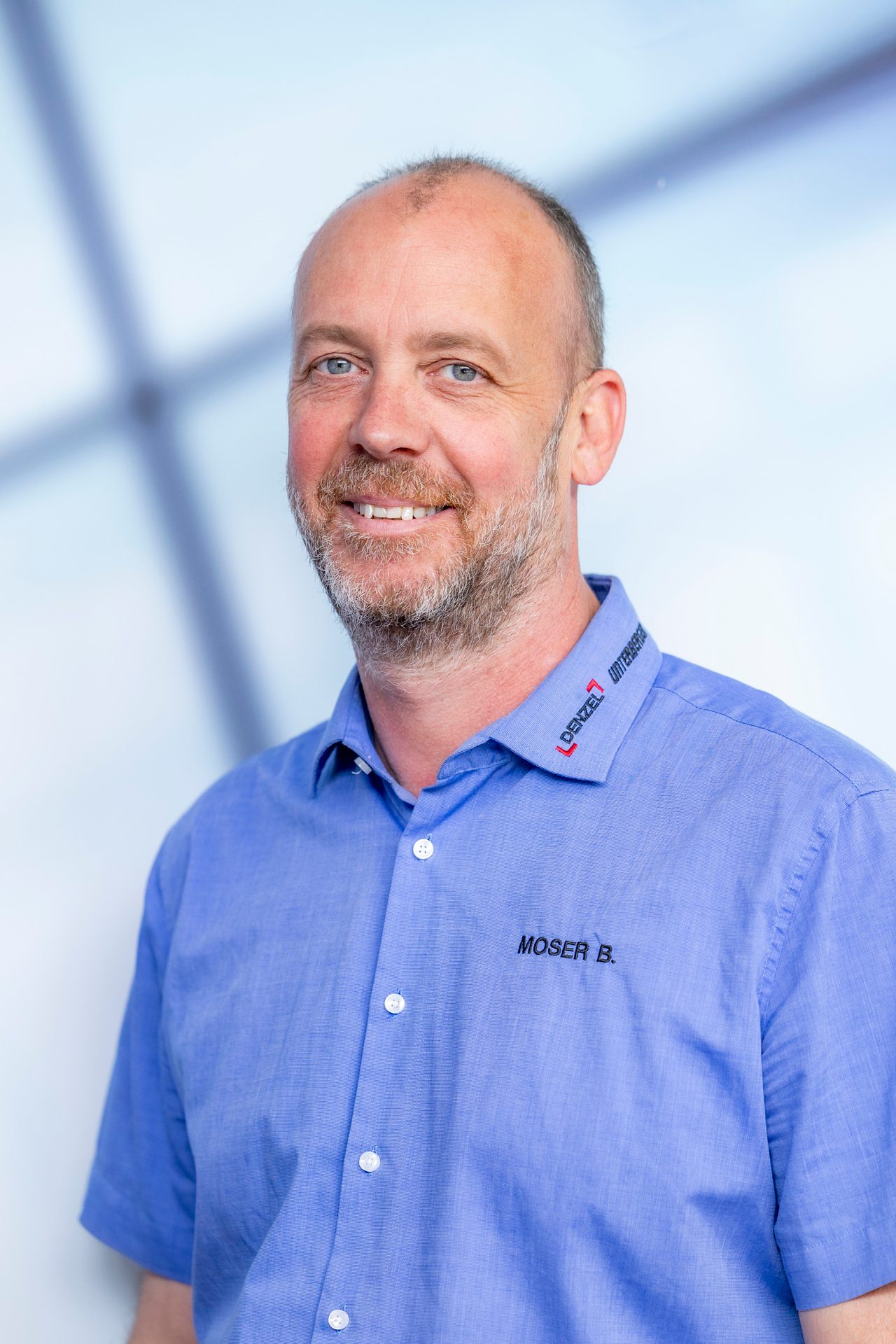 A smiling man with a beard and short hair, wearing a blue shirt with a company logo and a name tag.