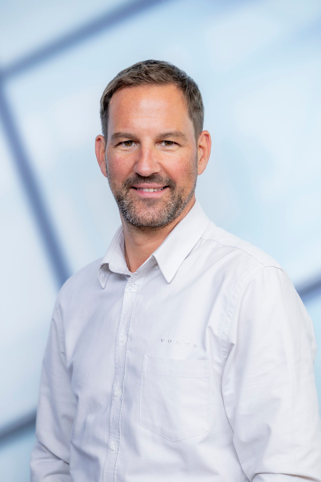 A smiling man with a beard and short hair, wearing a white Volvo shirt, looks at the camera.