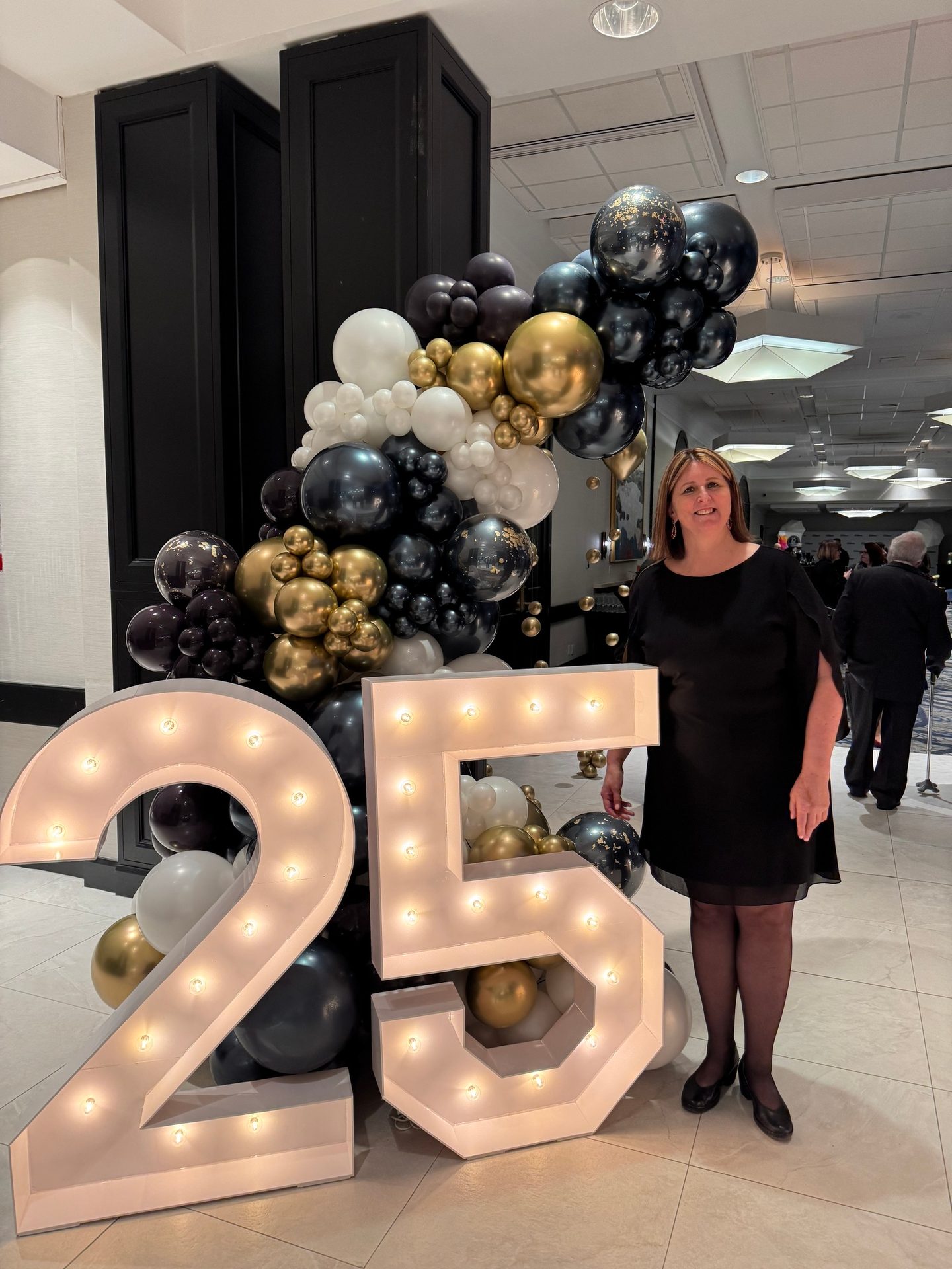 A smiling woman in a black dress stands next to large illuminated "25" numbers and a black, gold, and white balloon arch.