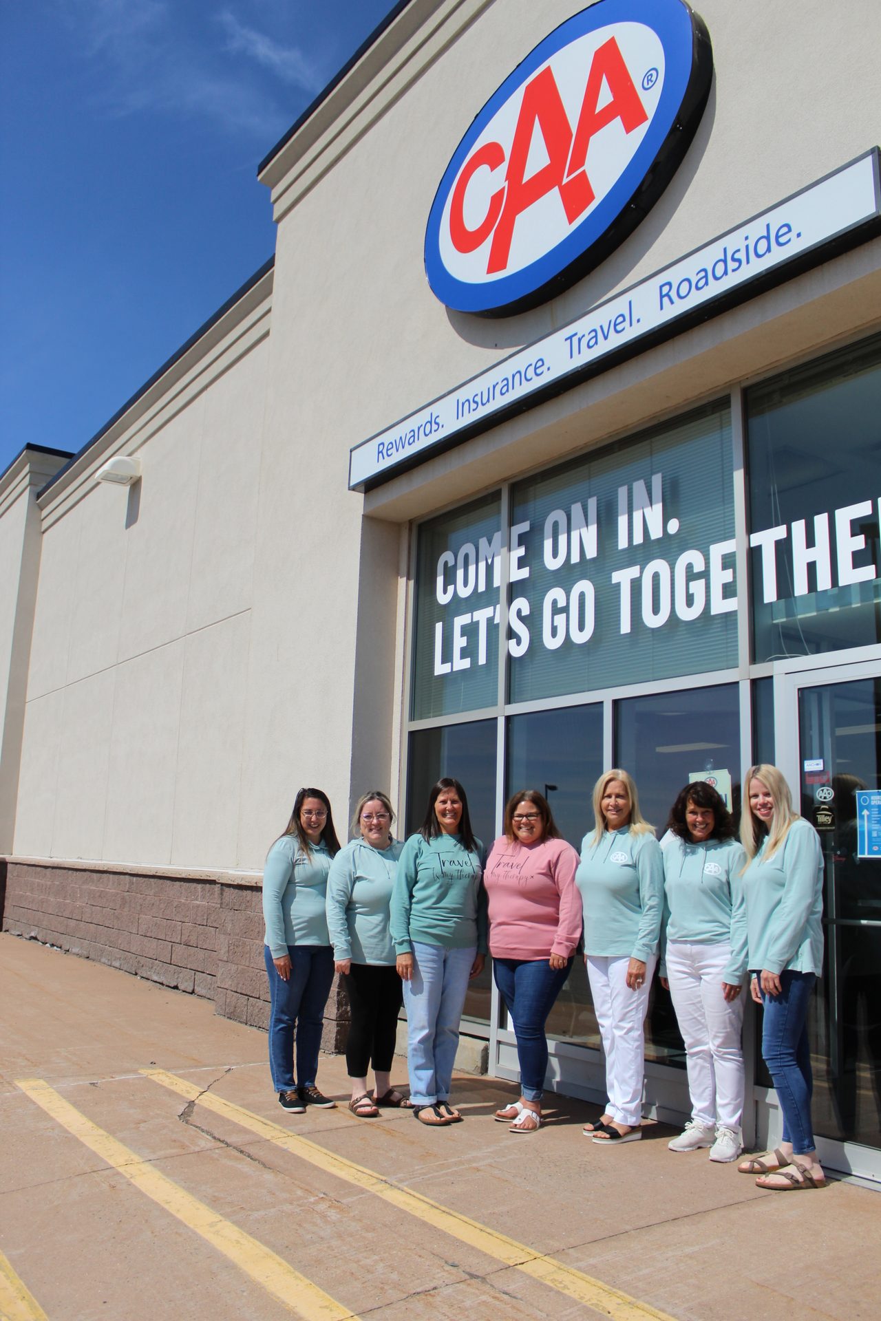 A group of women in light blue and pink tops standing in front of a CAA building under a clear blue sky.