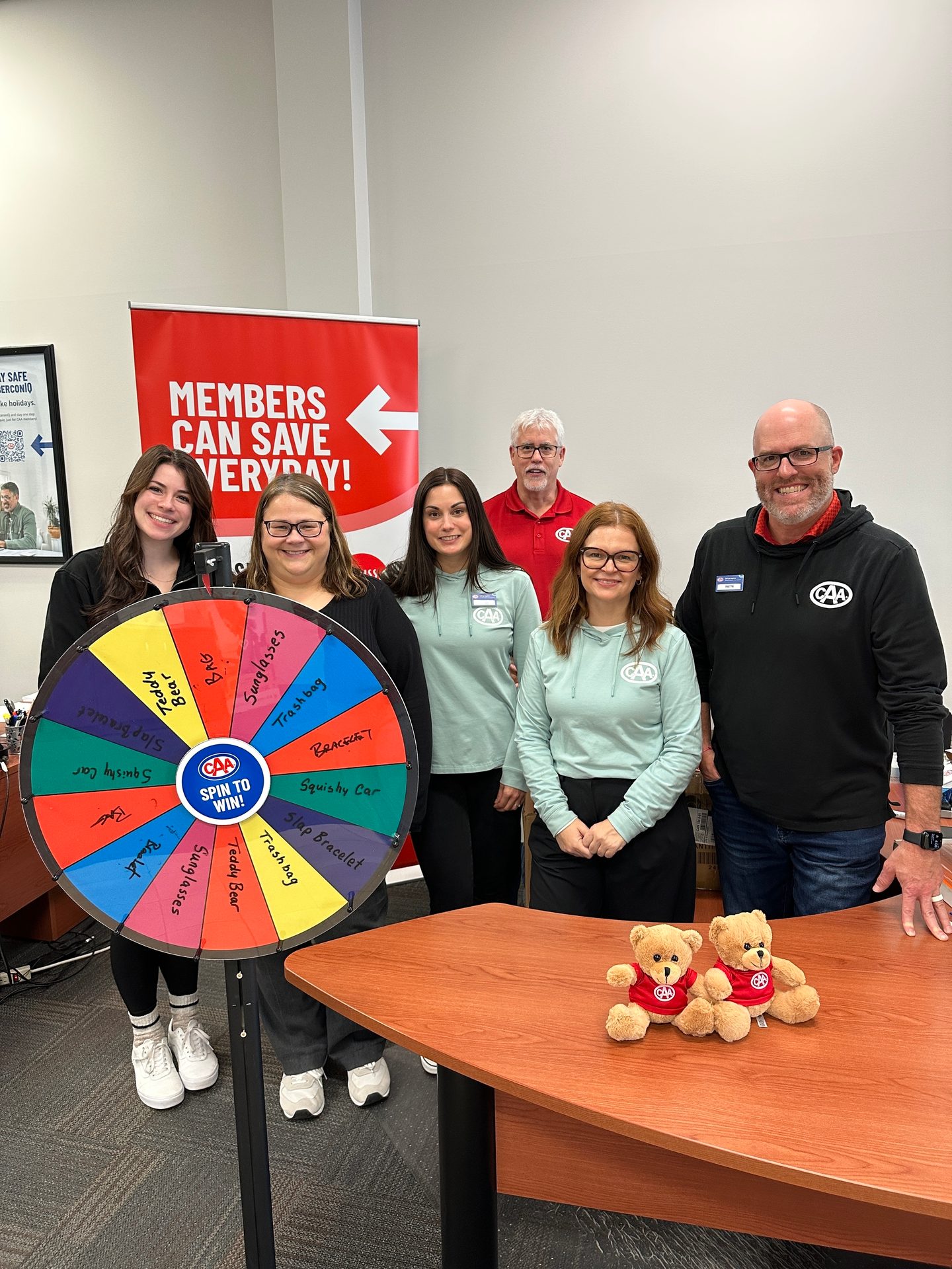 Six people pose with a "Spin To Win" wheel and two CAA teddy bears on a table.