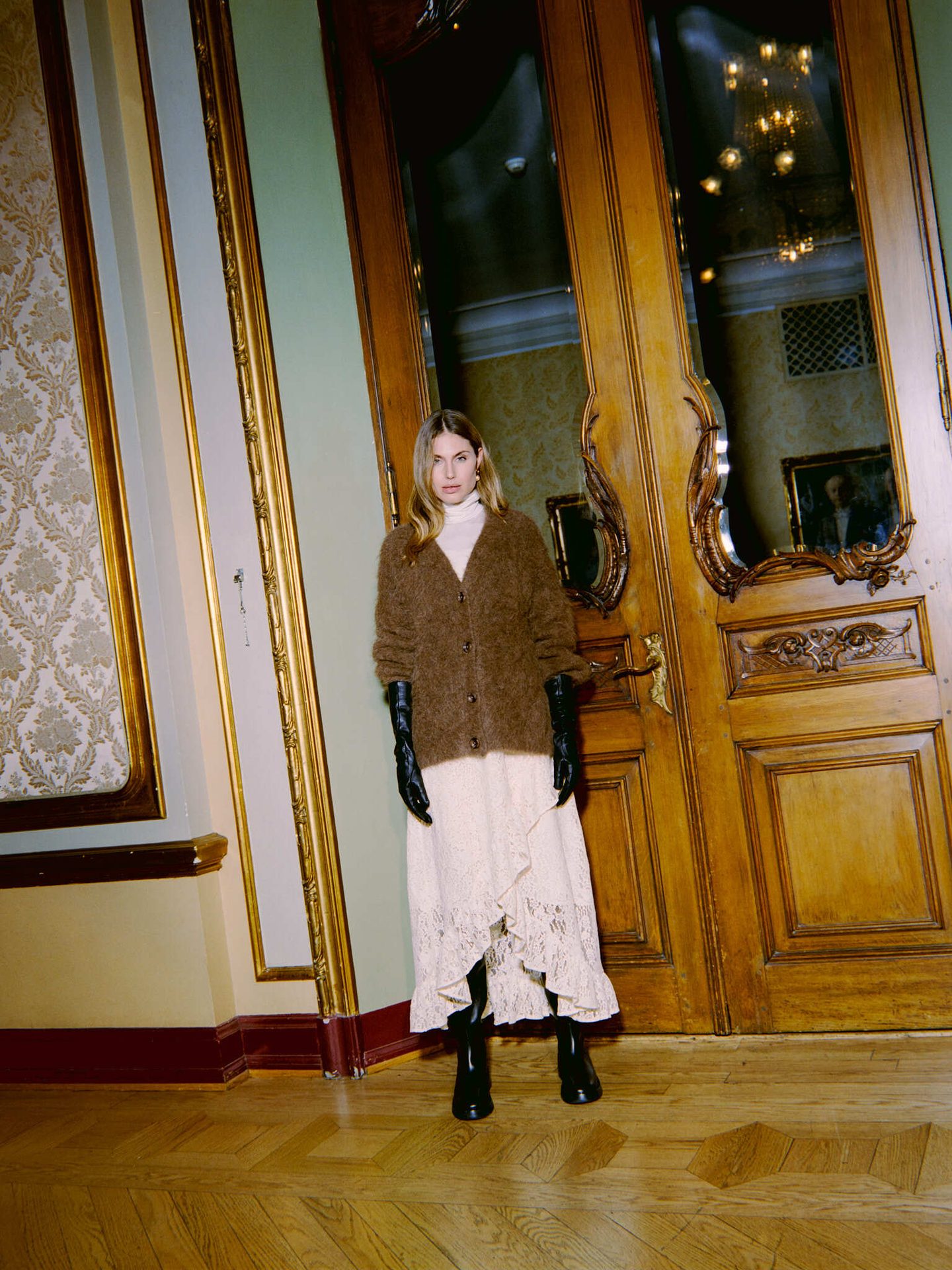 Woman in brown cardigan, cream skirt, and black boots, standing in ornate room with wooden doors.