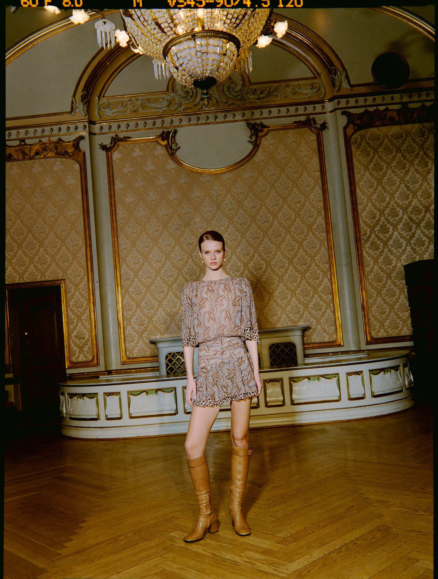 A woman in a patterned dress and brown boots stands in an elegant room with a chandelier.