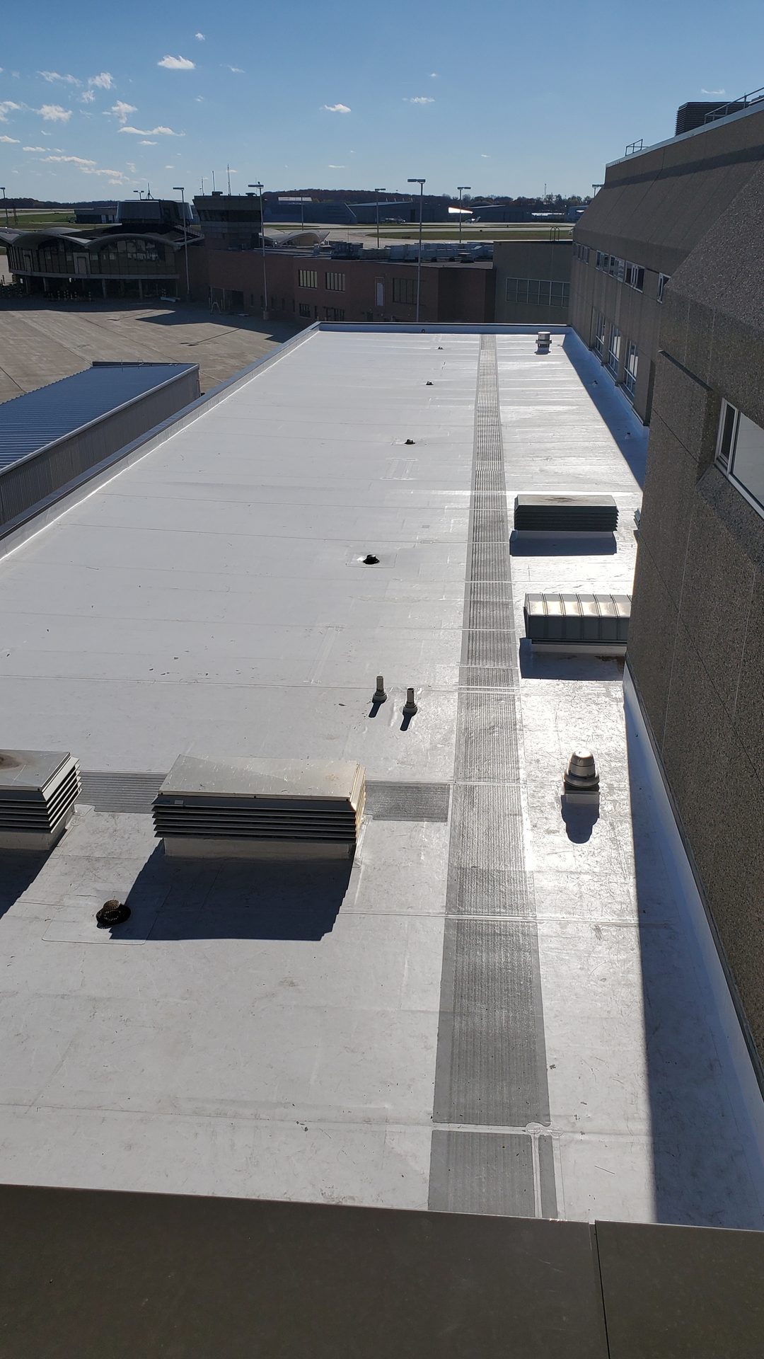 Aerial view of a large white flat commercial roof with HVAC units and a dark strip, overlooking an airport.