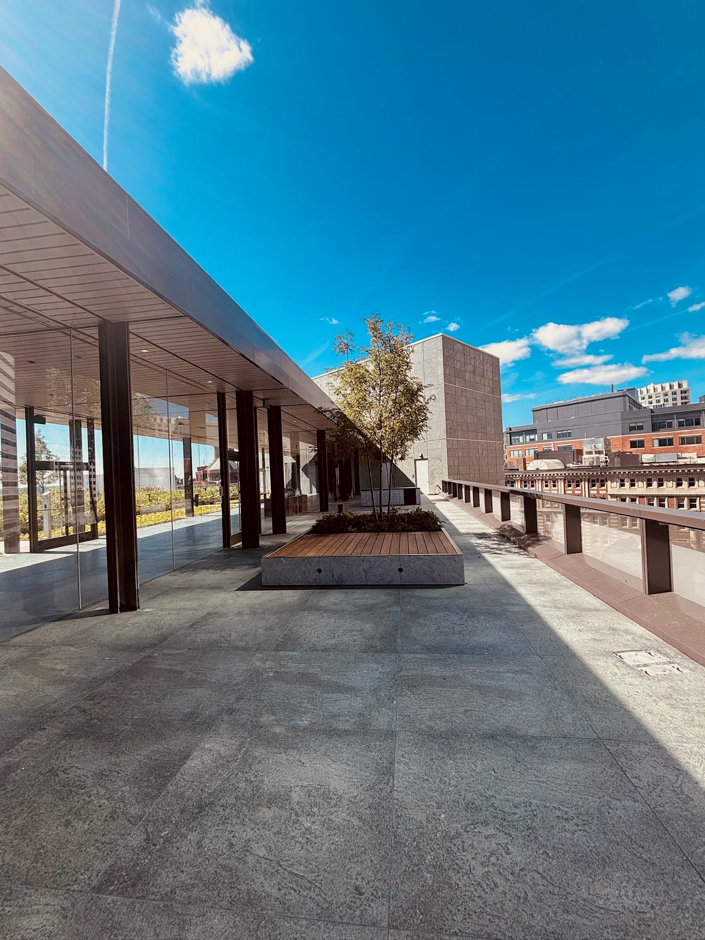 A modern rooftop terrace with a sleek building, large tiled floor, and a central planter under a blue sky.