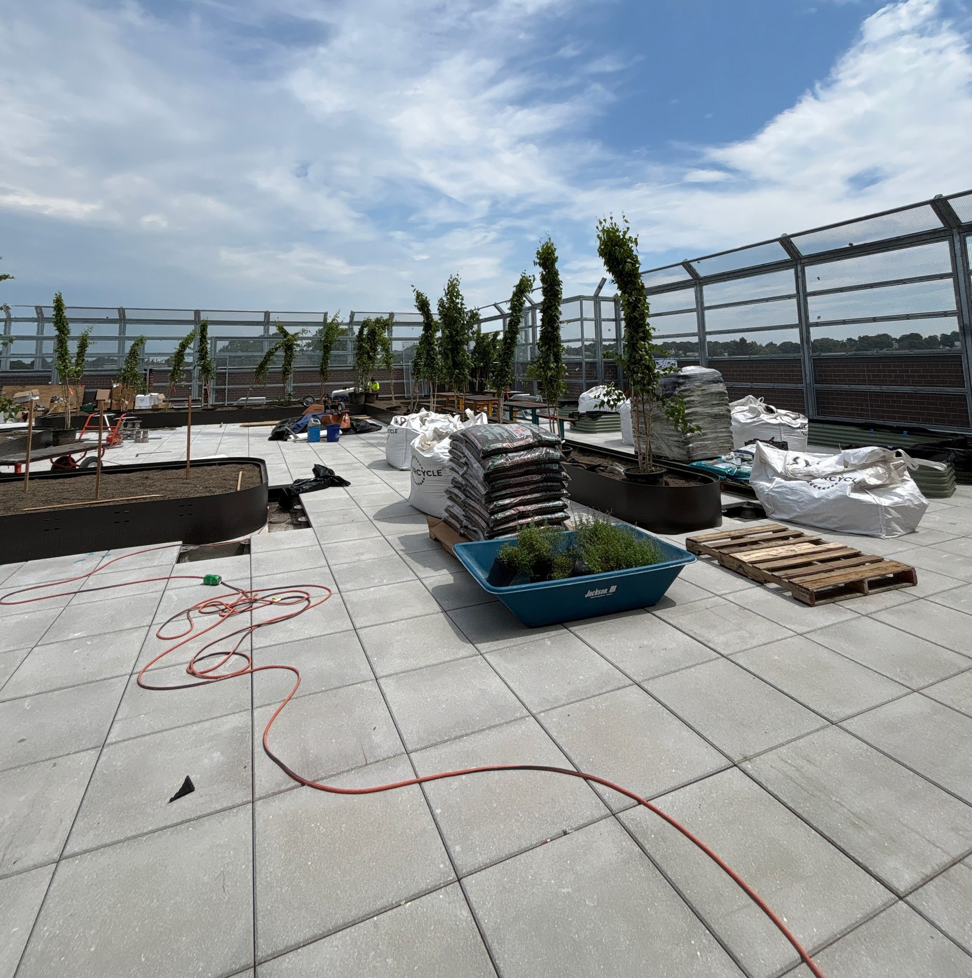 Rooftop garden under construction with trees in planters, bags of soil, plants in a wheelbarrow, and paving.