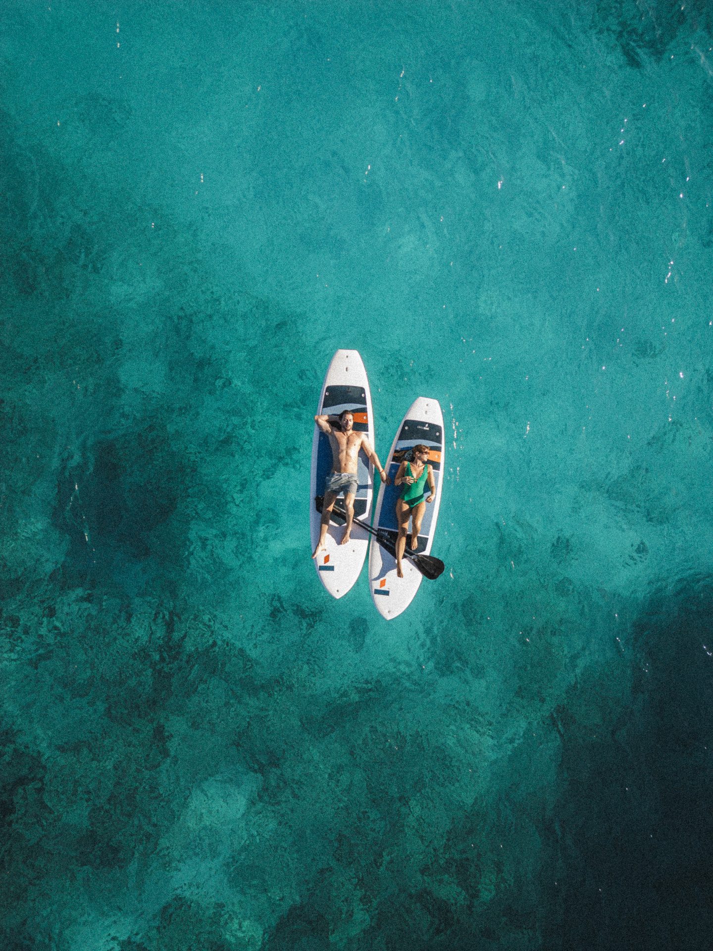 Aerial view of a man and a woman relaxing on paddleboards in clear turquoise water.