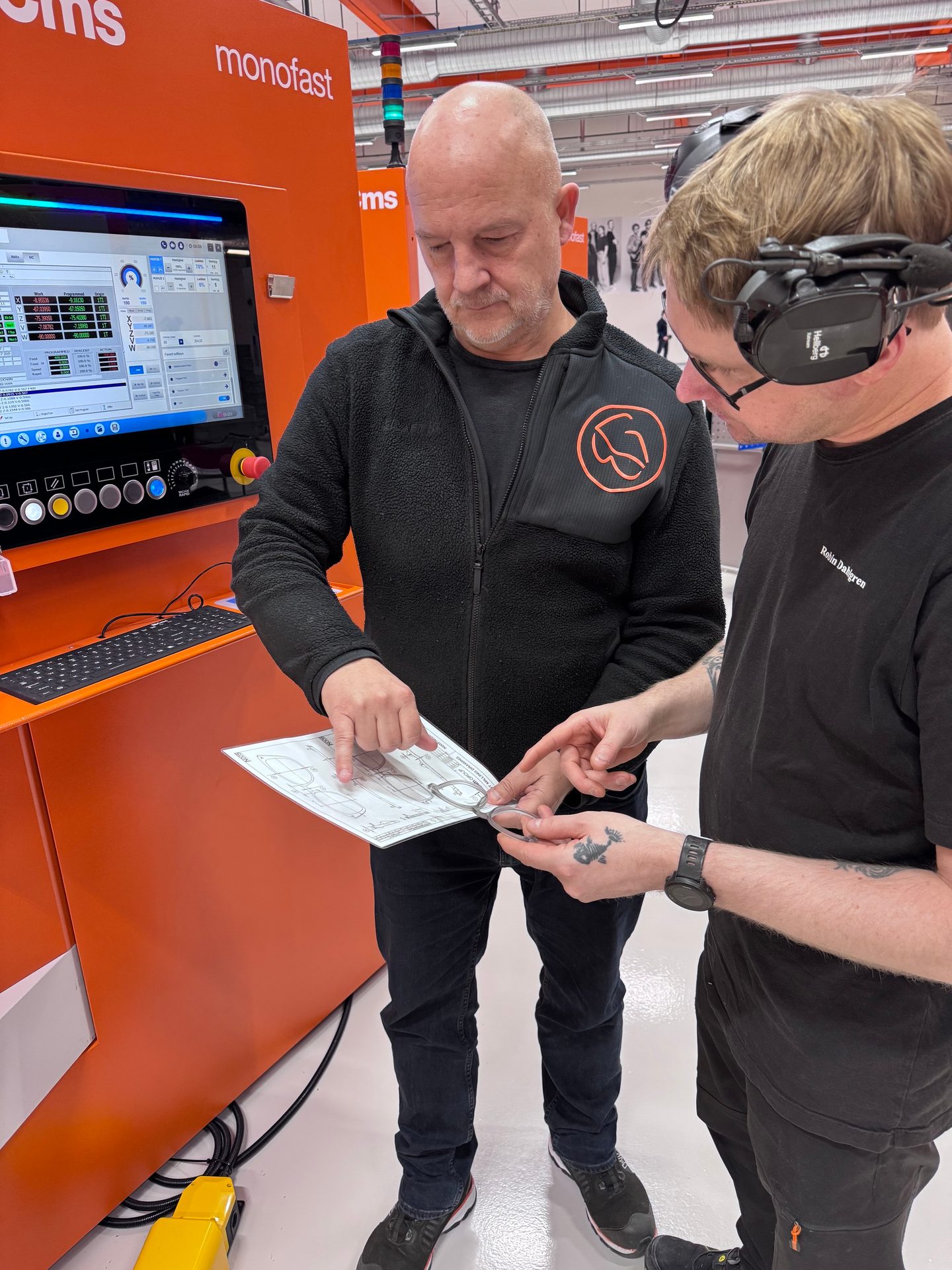 Two men in a workshop, looking at a diagram and a metal object by an orange machine.