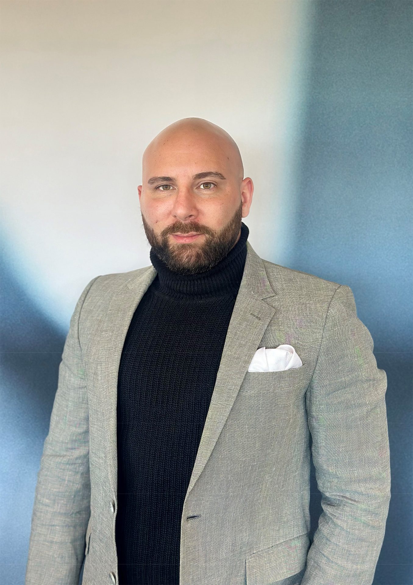 Bald man with beard in black turtleneck and grey blazer, looking forward.
