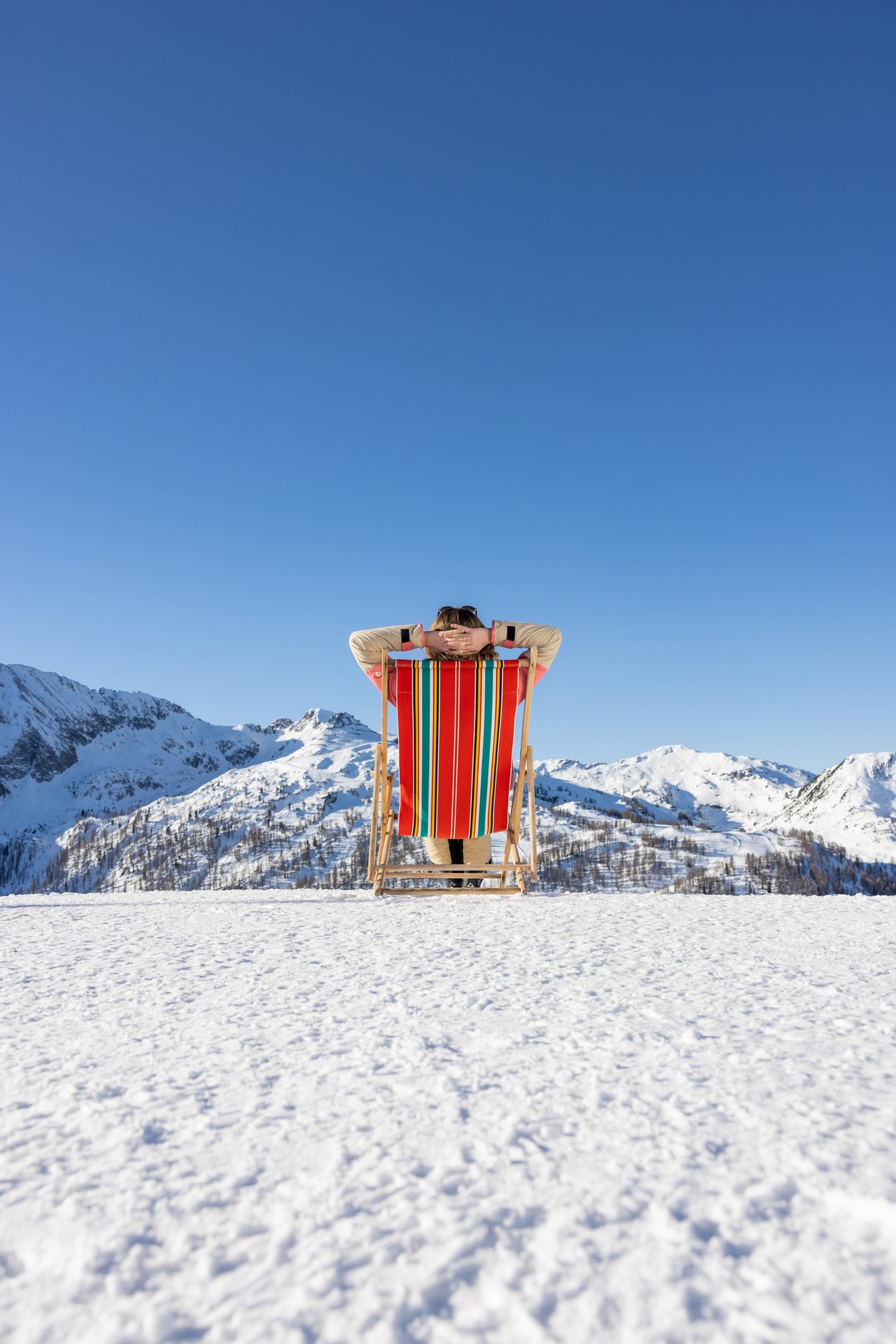 Person relaxing in a striped deck chair on a snowy mountain with clear blue sky and mountains.