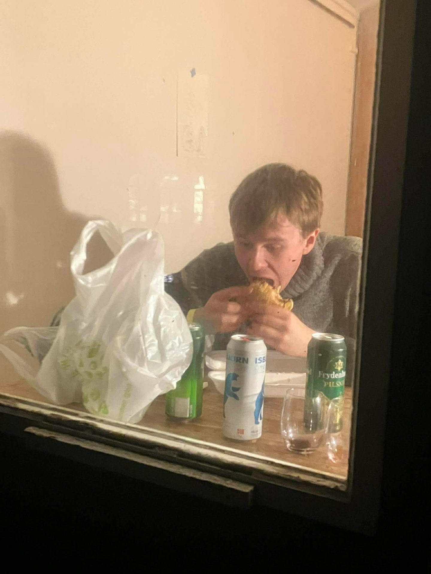 Young man intensely eating a large burger, viewed through a window, with drinks and a plastic bag on the table.
