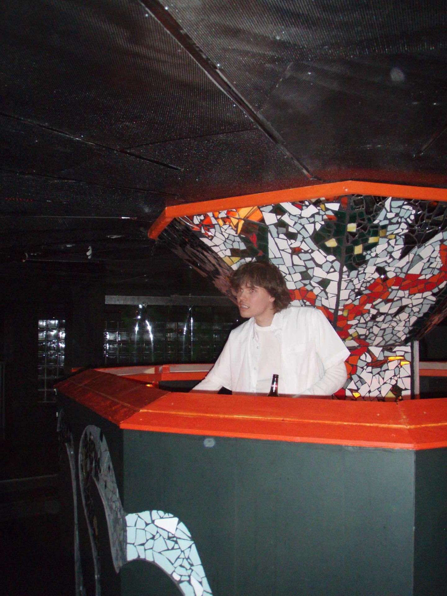 Young man in a white shirt behind a bar with colorful mosaic decorations, looking to the right.