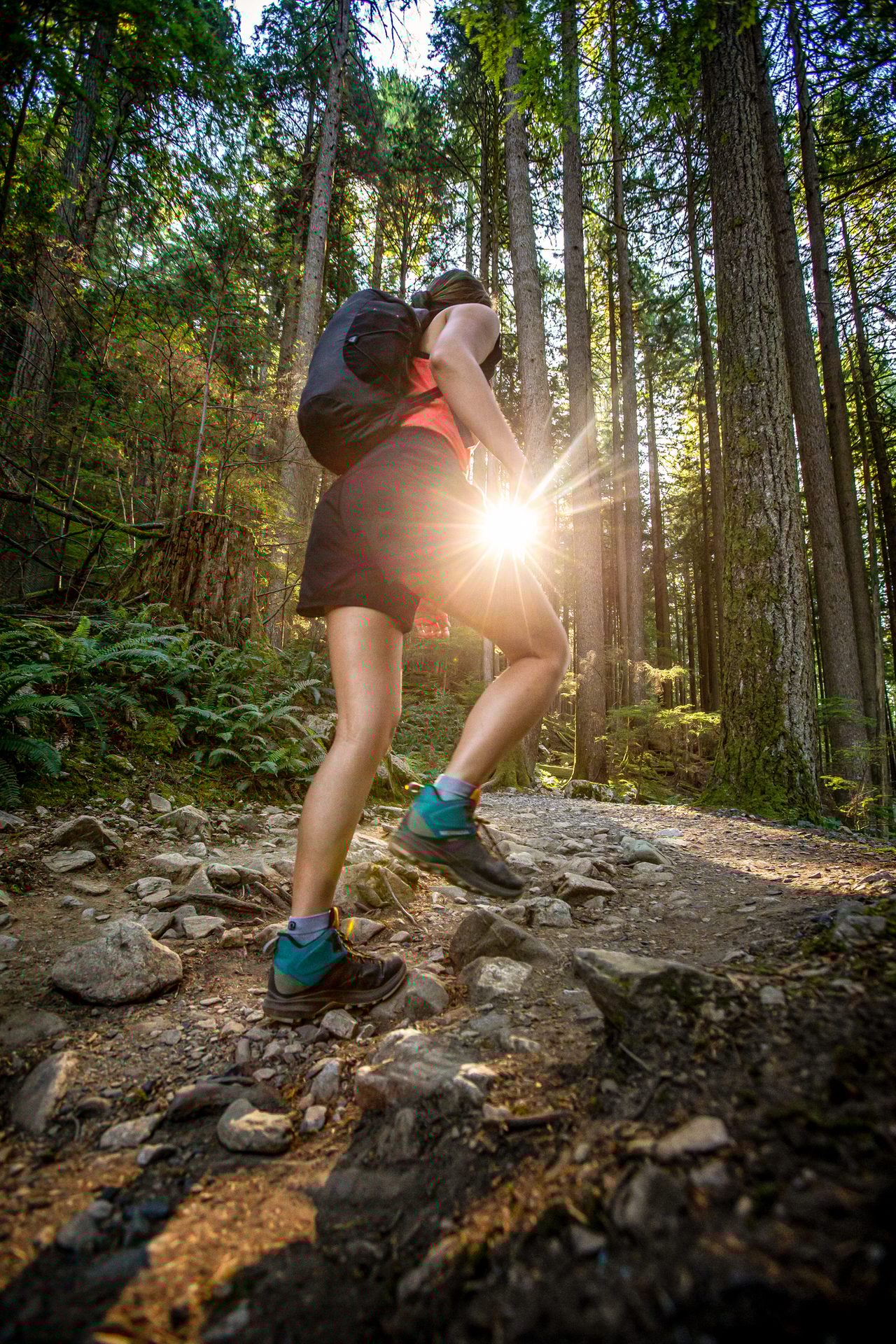 Hiker ascending a rocky forest path with sun flare, showing legs, boots, and backpack.