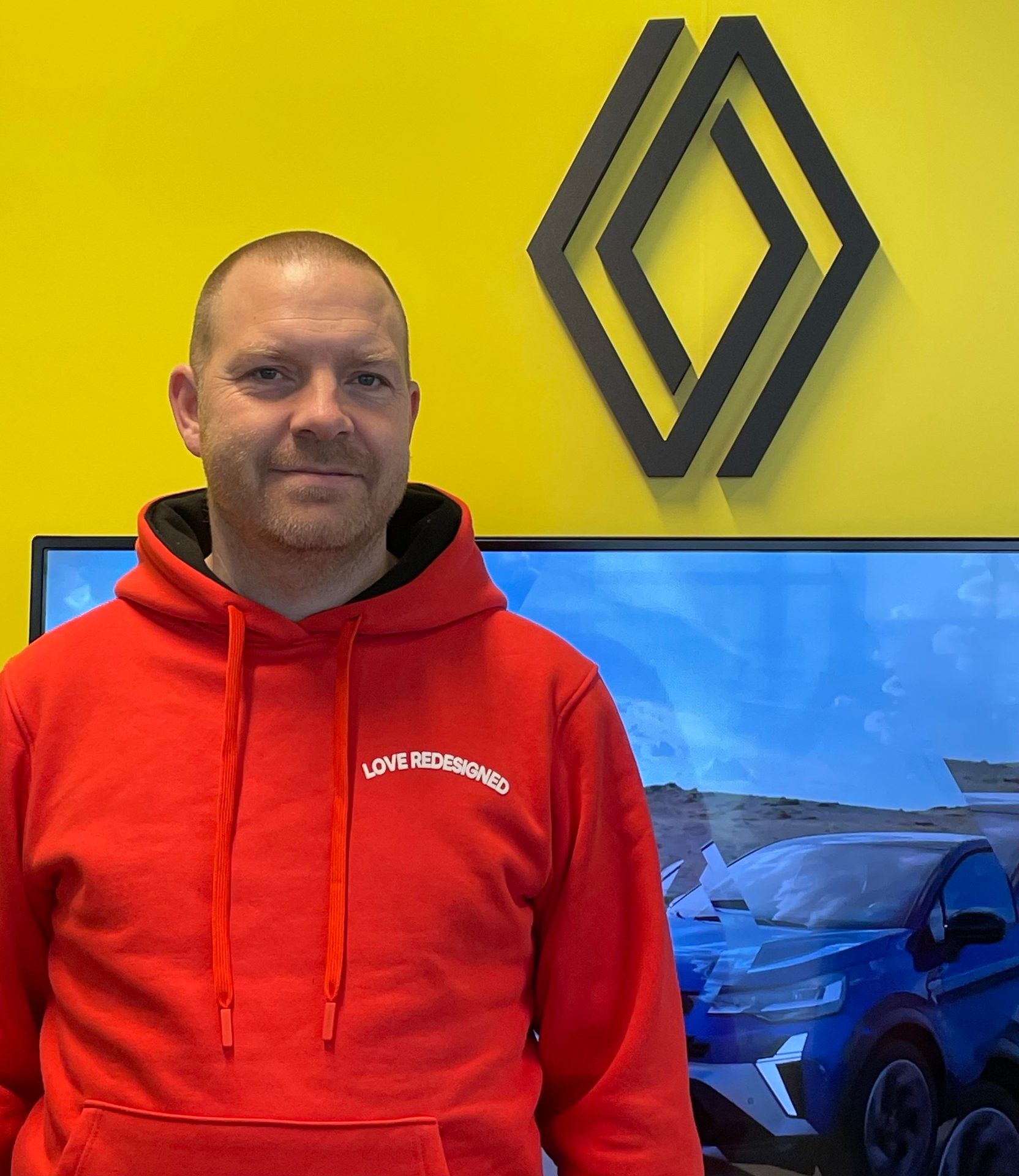 Man in a red 'LOVE REDESIGNED' hoodie against a yellow wall with a Renault logo and car on screen.